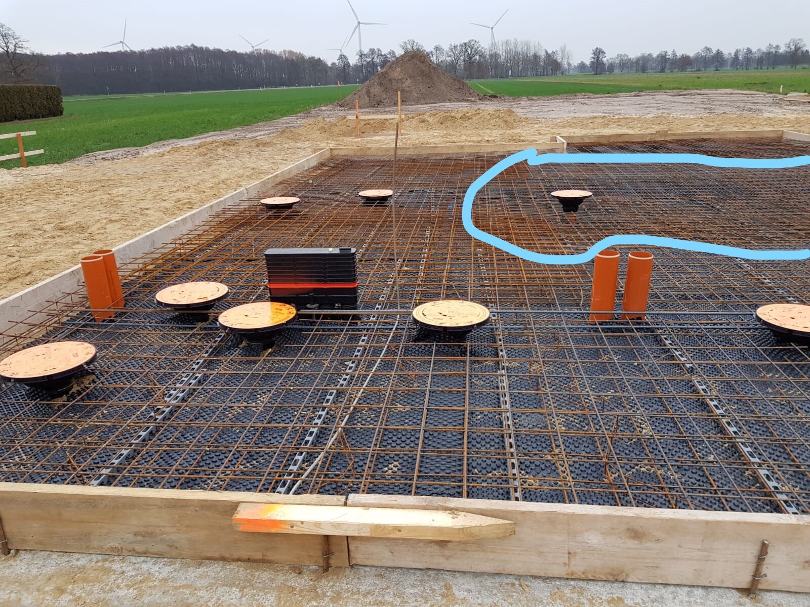 Fundamentfläche mit Bewehrungsstahlgitter, orange Rohre und Deckel, Windräder im Hintergrund.