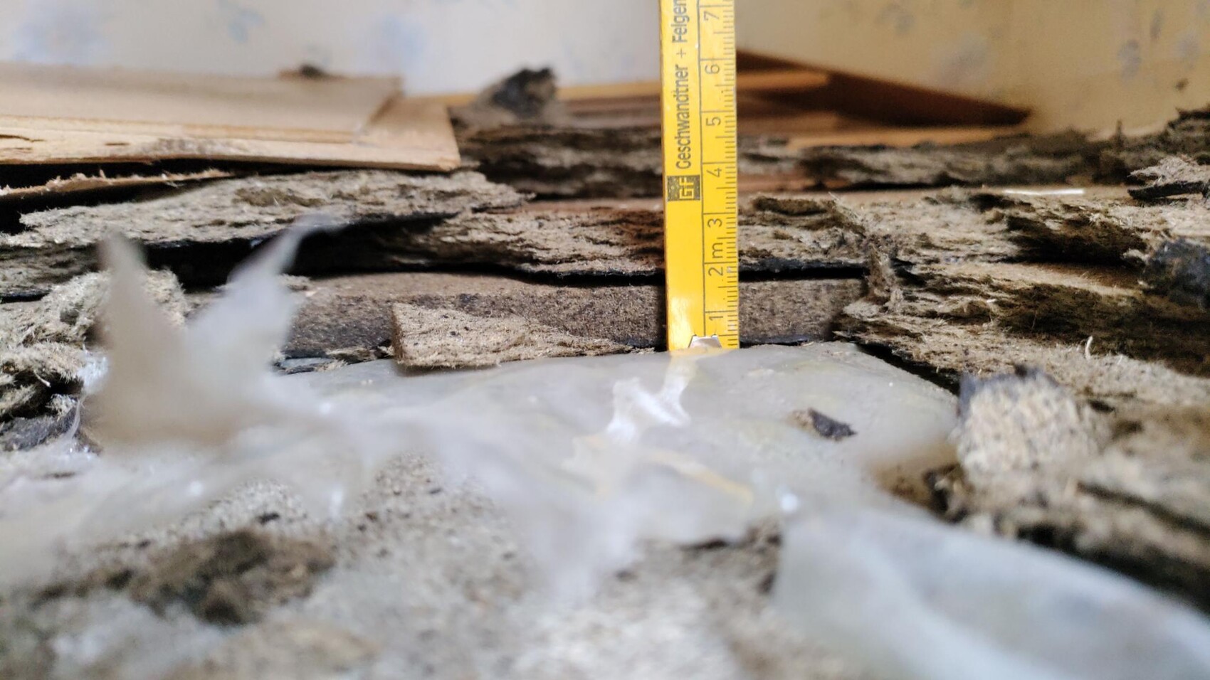 Nahaufnahme von beschädigtem Boden mit Holzplanken, Staub, Dämmmaterial und einem gelben Messband.