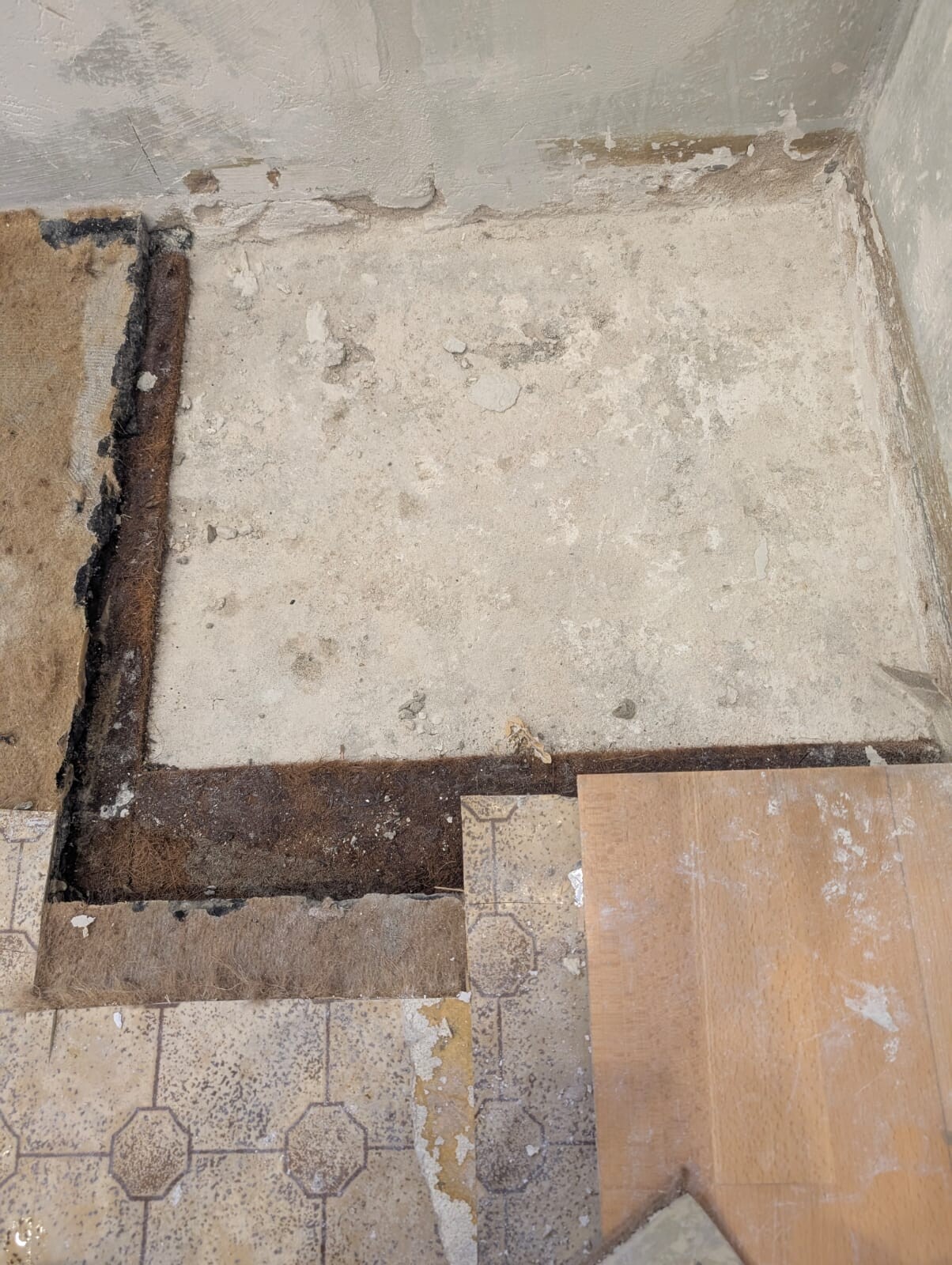 Bodenöffnung in altem Raum: rostiger Stahlrahmen, bröckelnde Fliesen und eine Holzplatte.