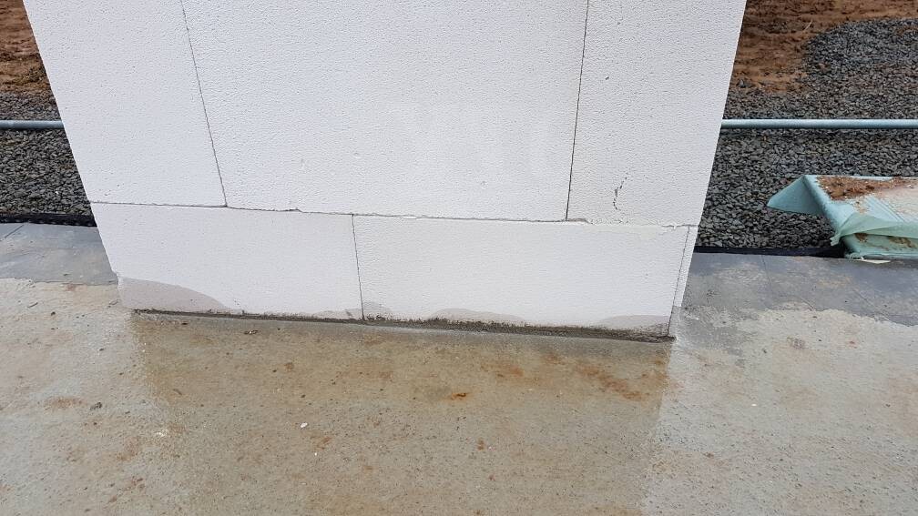 Weiße Betonblockwand auf nassem Betonboden; Feuchtigkeit, Rohr und Baumaterial am Rand sichtbar.