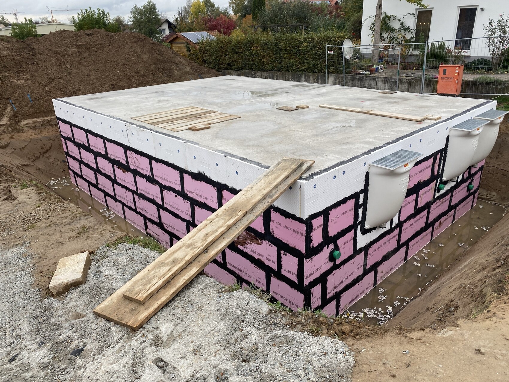 Betonfundament eines Gebäudes mit pinker Isolierung, schwarze Fugen, Holzplanken und Rohre.