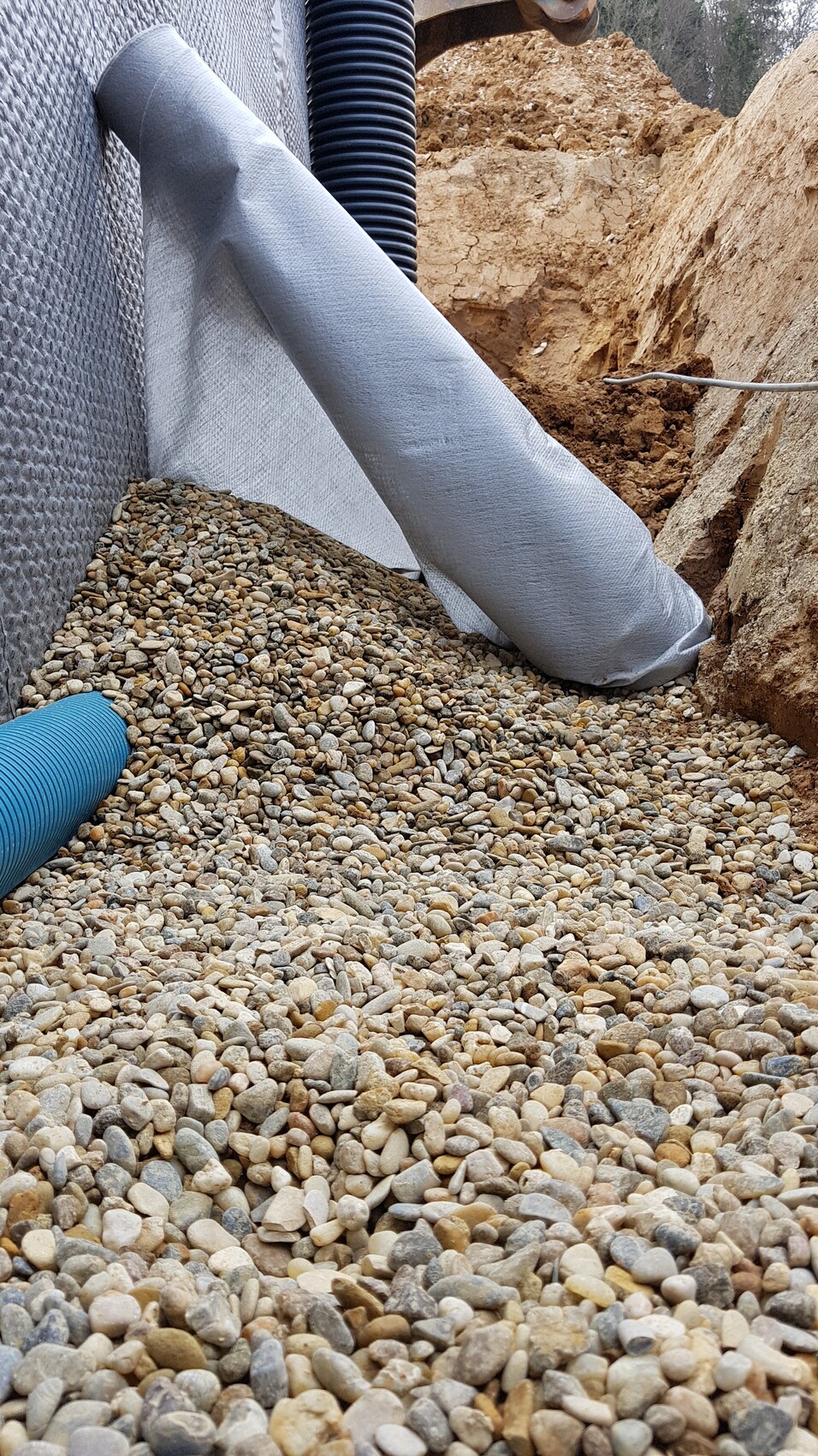 Graben auf Baustelle mit Kiesbett, Drainagerohr und blauem Schlauch; Geotextil liegt bereit.