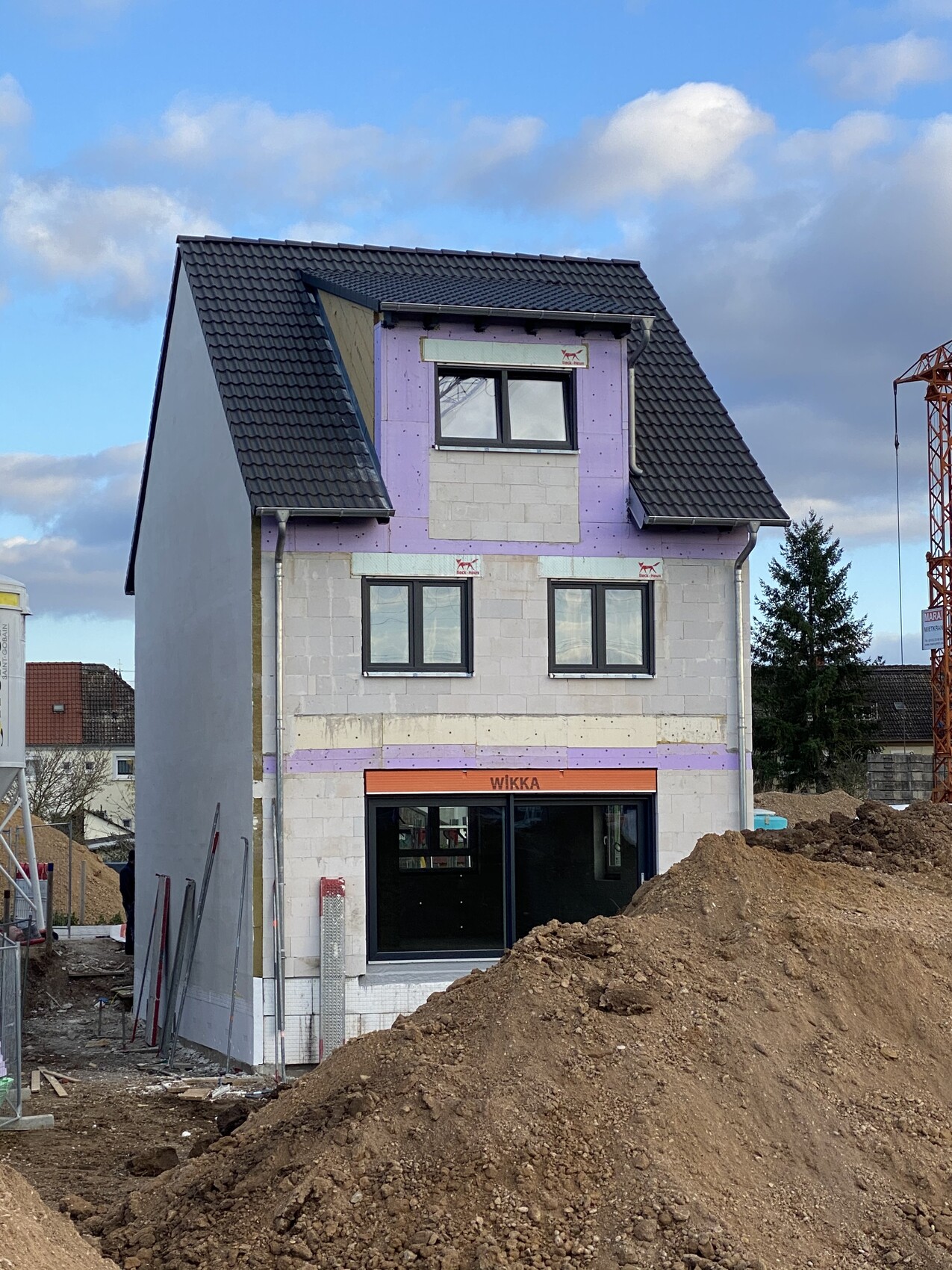 Unvollständiges Haus im Bau mit violetter Dämmung, Erdhaufen und Kran im Hintergrund