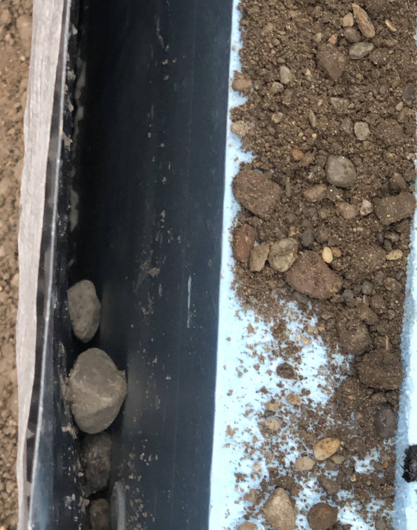 Nahaufnahme eines schwarzen Metallrahmens im Bau mit Erde und Kies neben einer hellblauen Fläche.