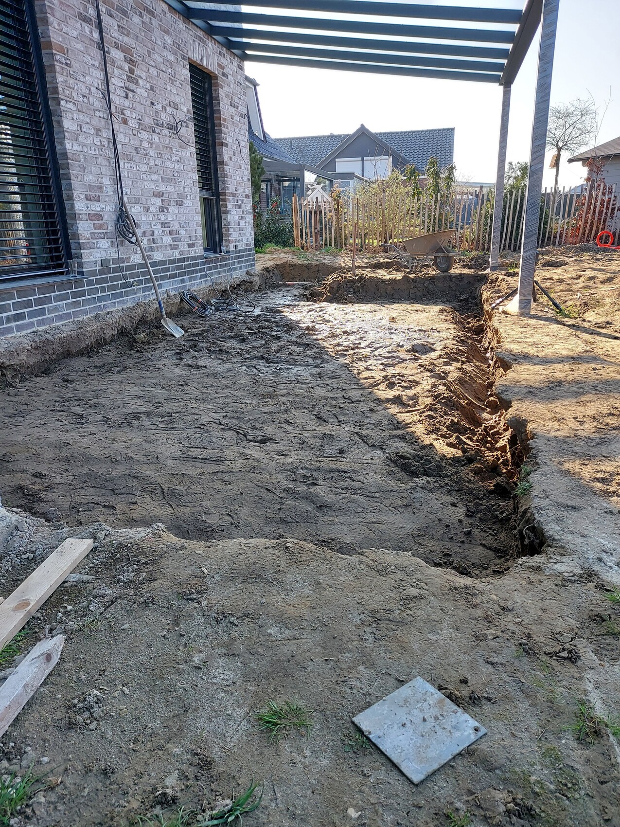 Ausgehobene Baugrube im Hinterhof neben einem Haus, Schaufel und Holzplanken liegen daneben.