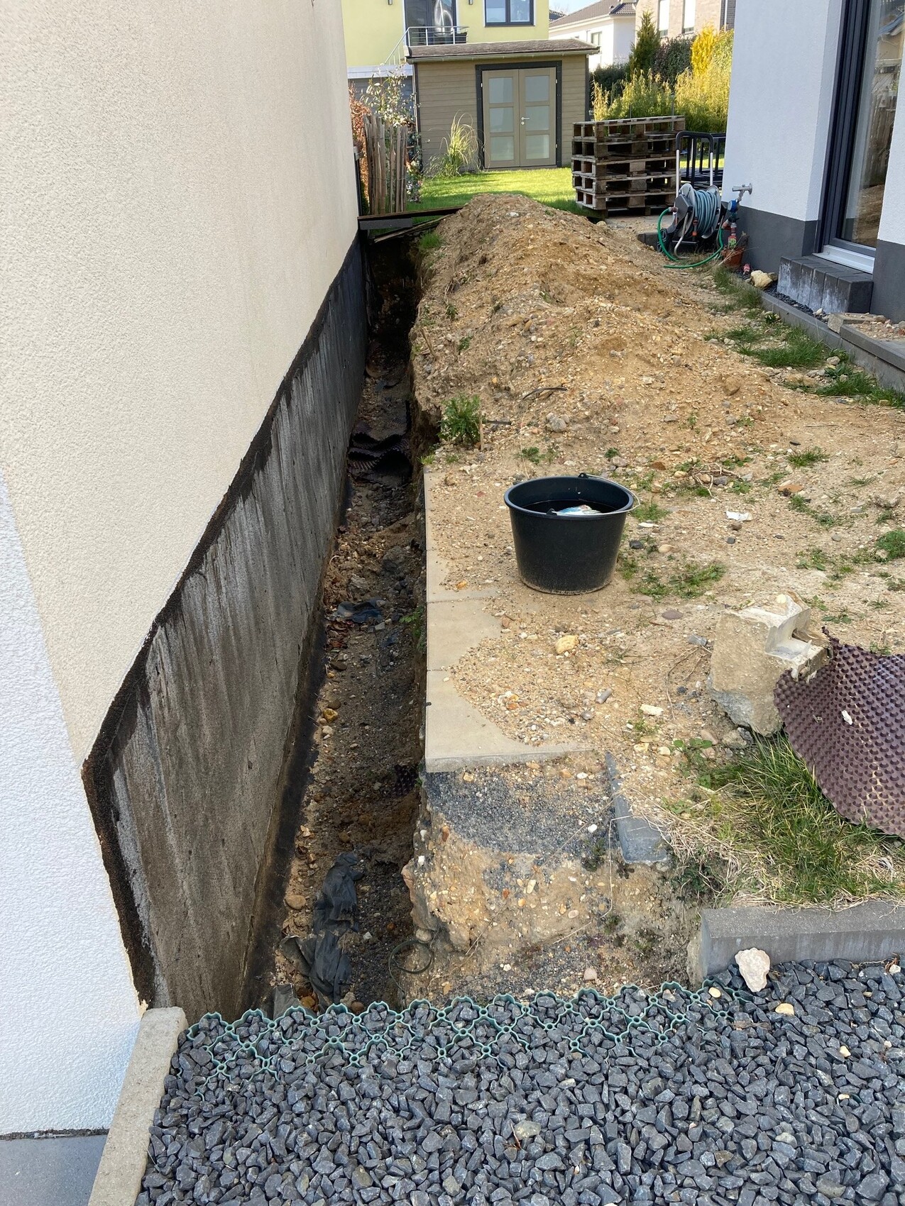Graben neben Hauswand mit Erdaushub, Eimer, Schlauchaufroller und Paletten im Garten