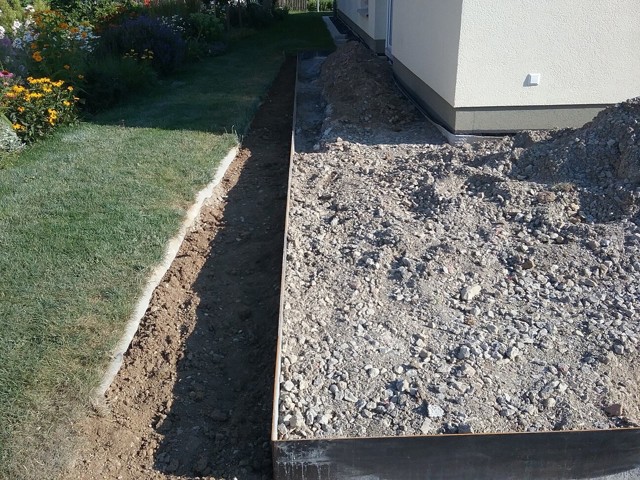 Baugrube entlang der Hauswand mit Metallrand, Kieshaufen rechts, Rasen und Blumen links.