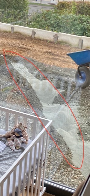 Baugrube mit Schlamm, blaue Schubkarre rechts, Metallgeländer im Vordergrund.