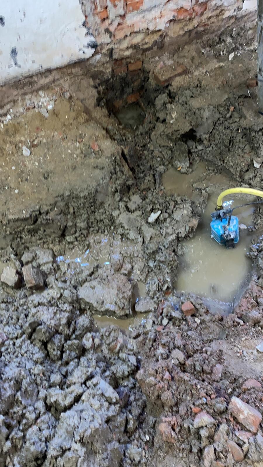 Baugrube mit schlammigem Boden, freigelegte Ziegelwand und blaue Sumpfpumpe mit gelbem Schlauch.