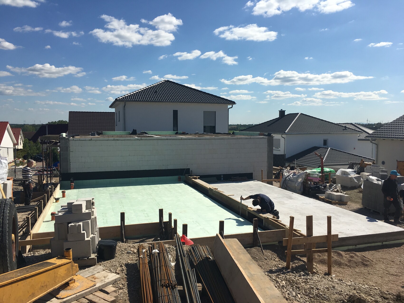 Unfertige Baustelle mit grünem Betonboden, Steinen, Holzplanken und Arbeitern vor Häusern.