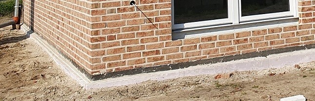 Eckansicht eines Backsteinhauses mit Fenster; Boden und Fundamentbereich mit Erde.