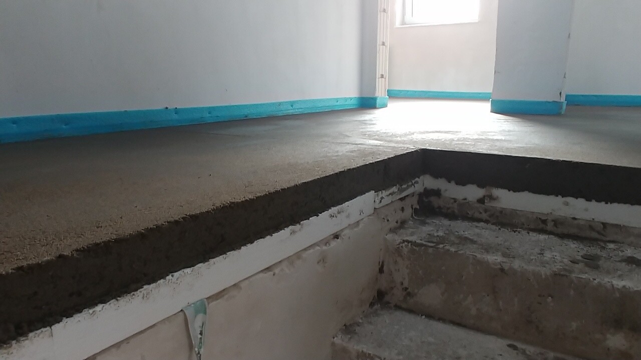 Innenraum einer Baustelle: roher Betonboden, Graben, blaue Sockelleisten, Licht durch Tür.