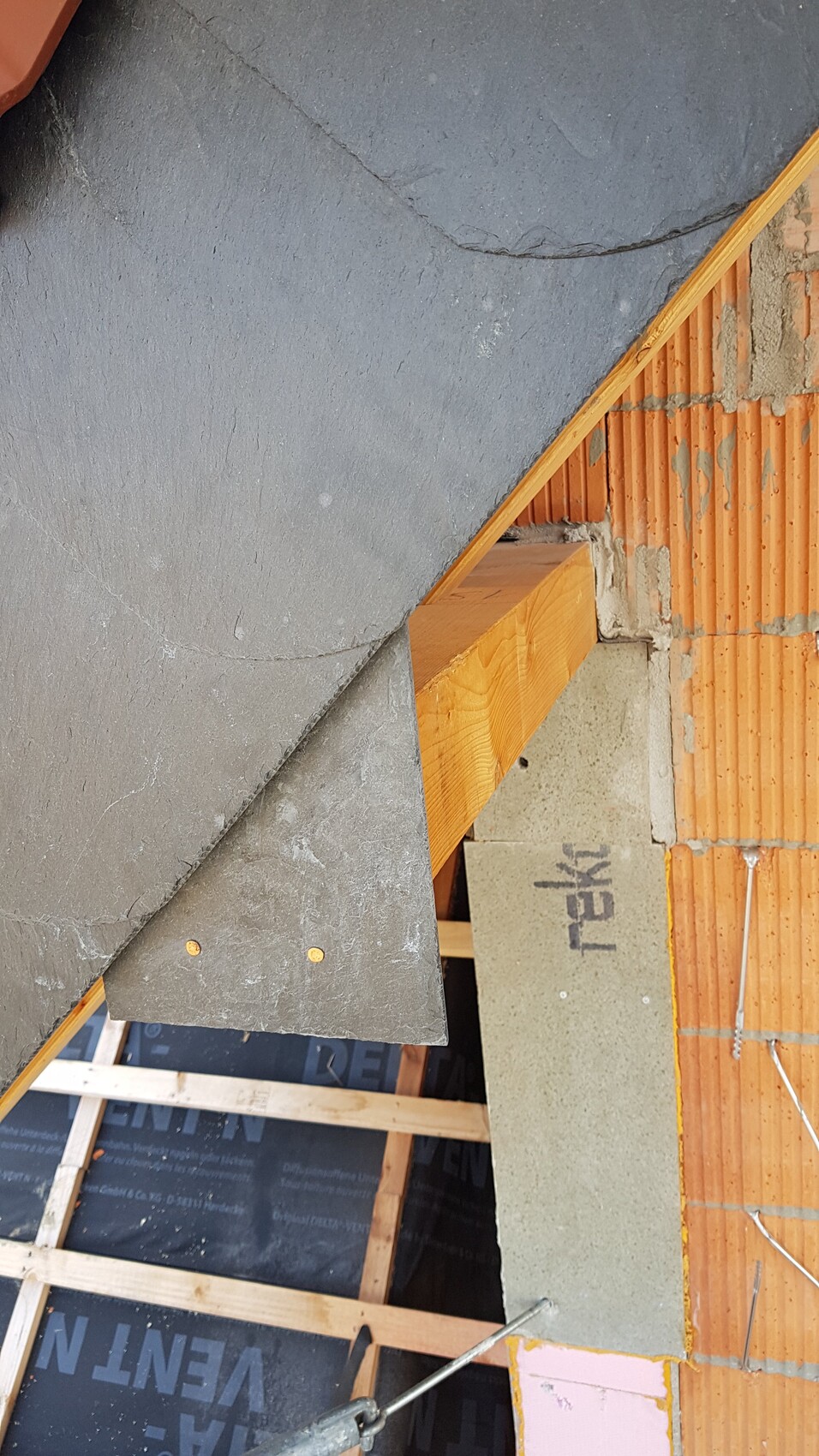 Rohbaustelle: graue Dachplatten, Holzbalken, rote Ziegelwand, Betonblock, Baufolie am Boden.