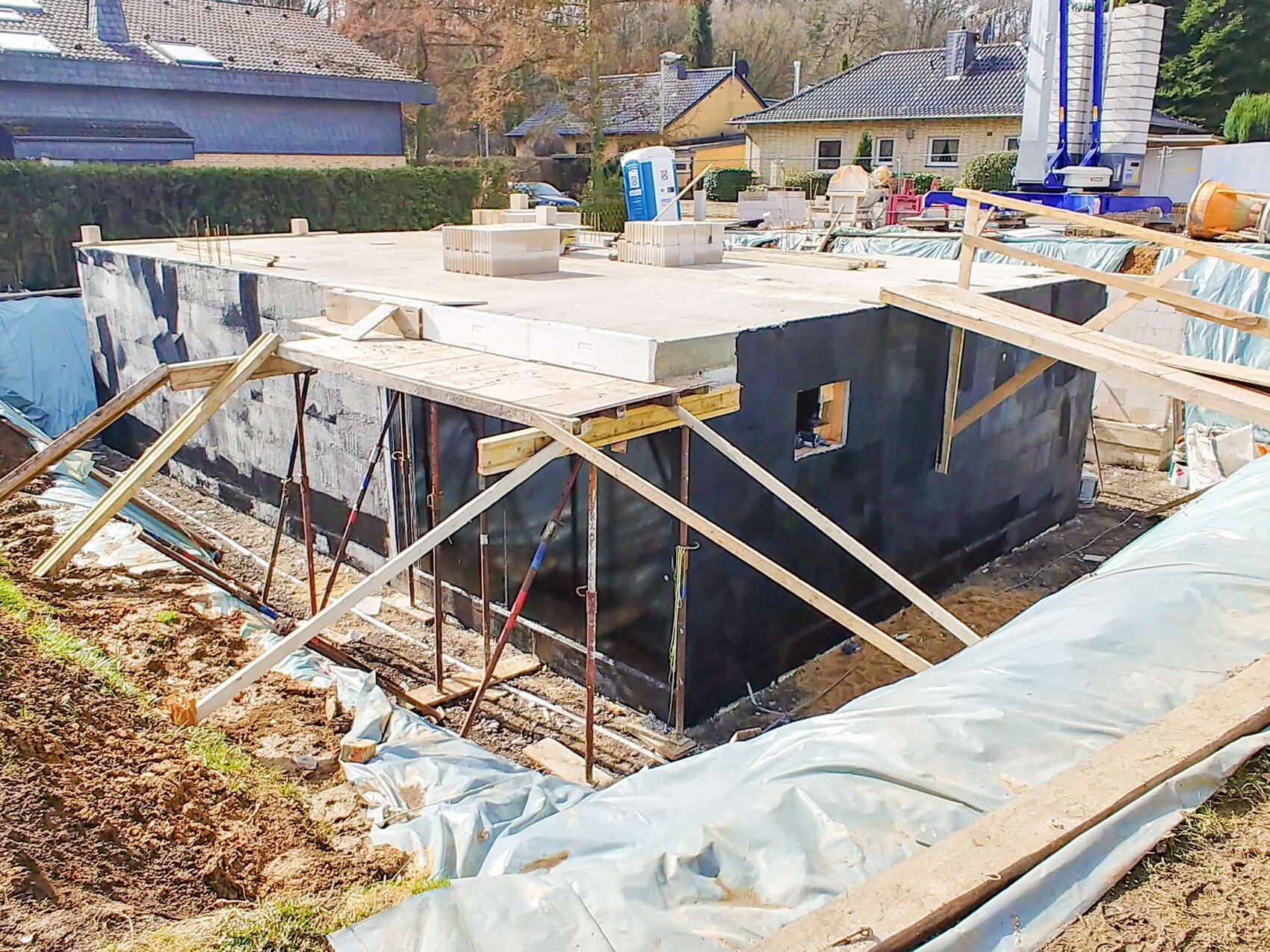 Baugrube mit Fundamentplatte und dunkler Abdichtung, Holzstützen, Häuser im Hintergrund