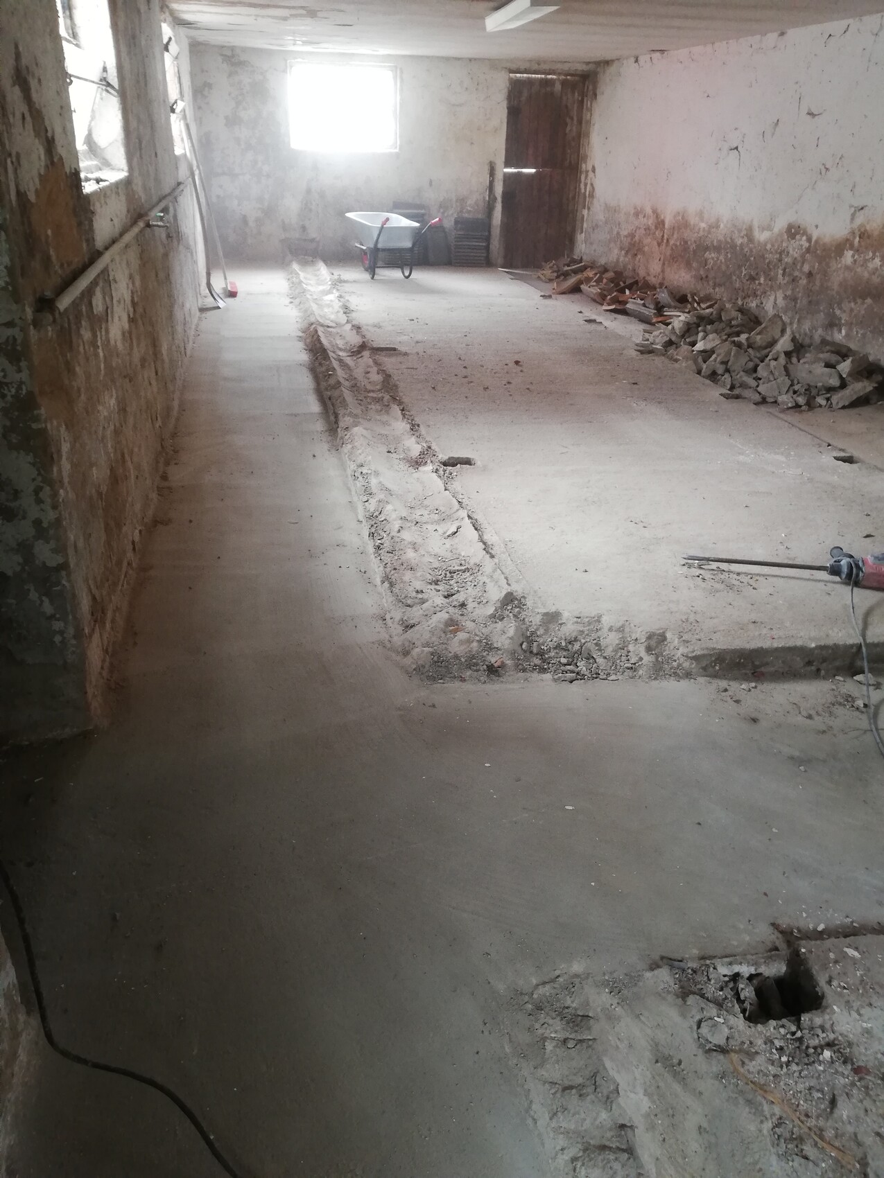Kellerbaustelle: rauer Betonboden, Graben in der Mitte, Schutt rechts, Wheelbarrow im Hintergrund.