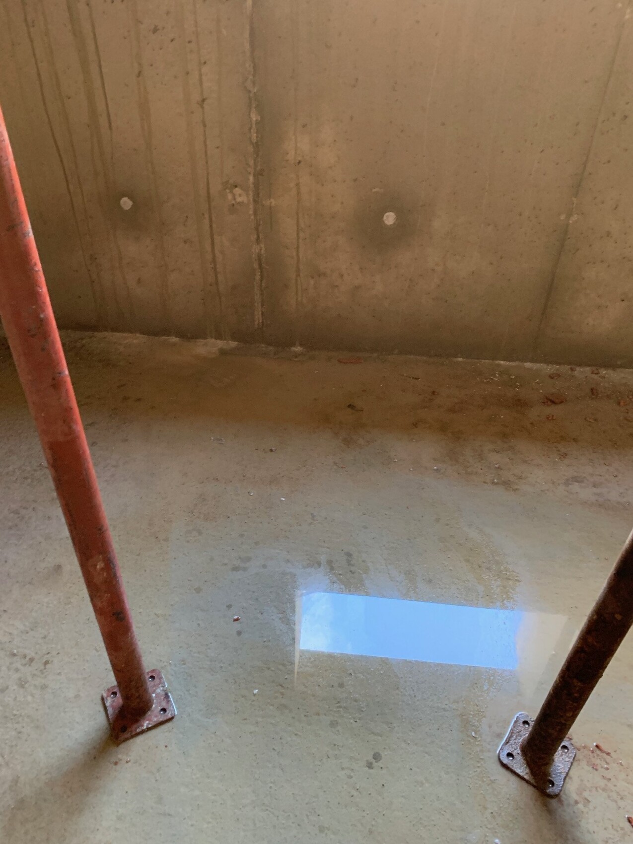 Zwei rostige Stahlstützen stehen auf Betonboden; eine Pfütze spiegelt ein blaues Rechteck.