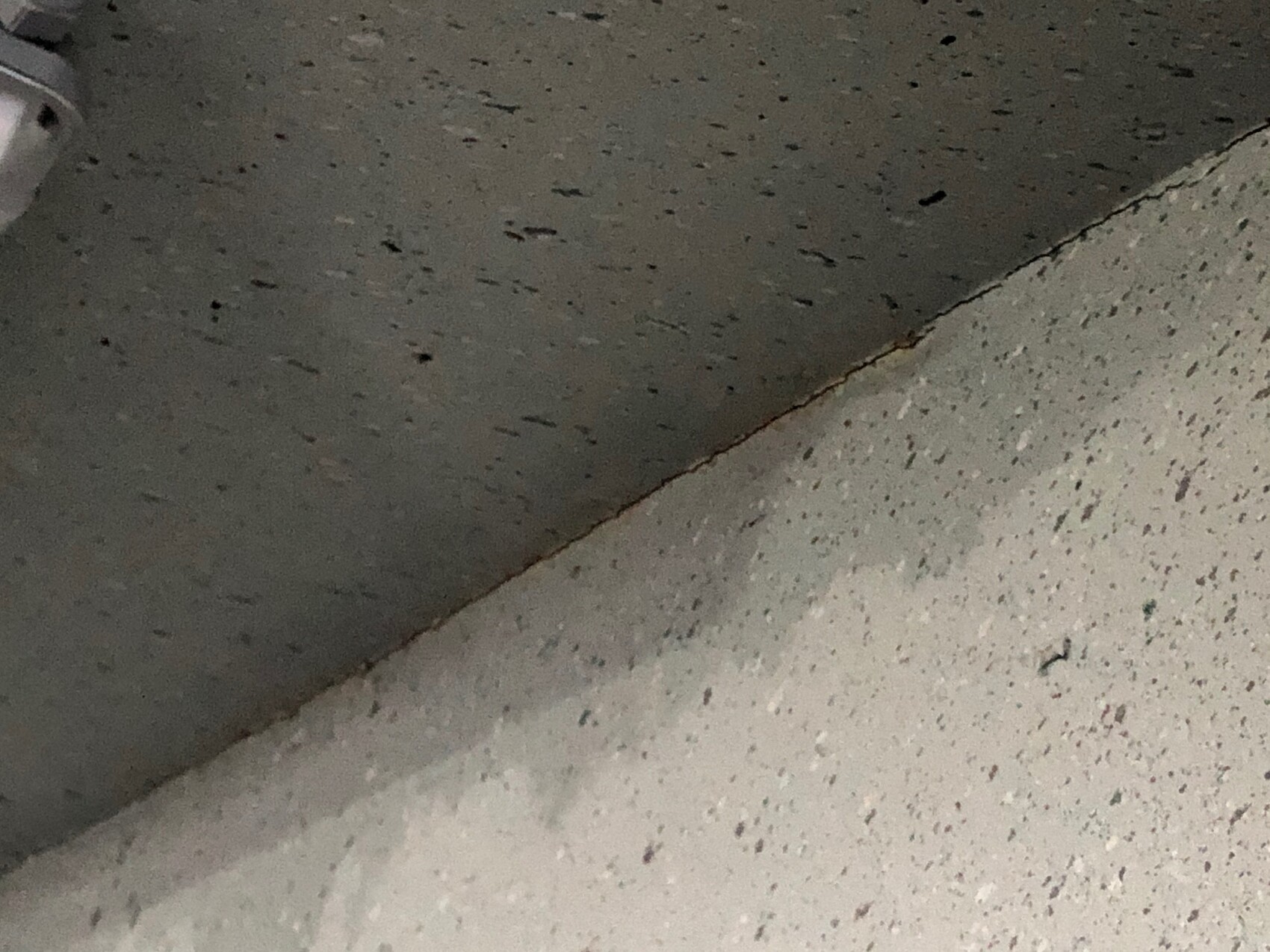 Nahaufnahme einer Ecke: dunkle obere Fläche trifft diagonal auf hellgrauen Boden mit kleinen Flecken.