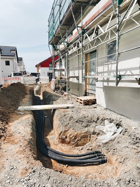 Baugrube mit schwarzen Rohren neben Gebäude, Holzbrücke über Grube, Gerüst