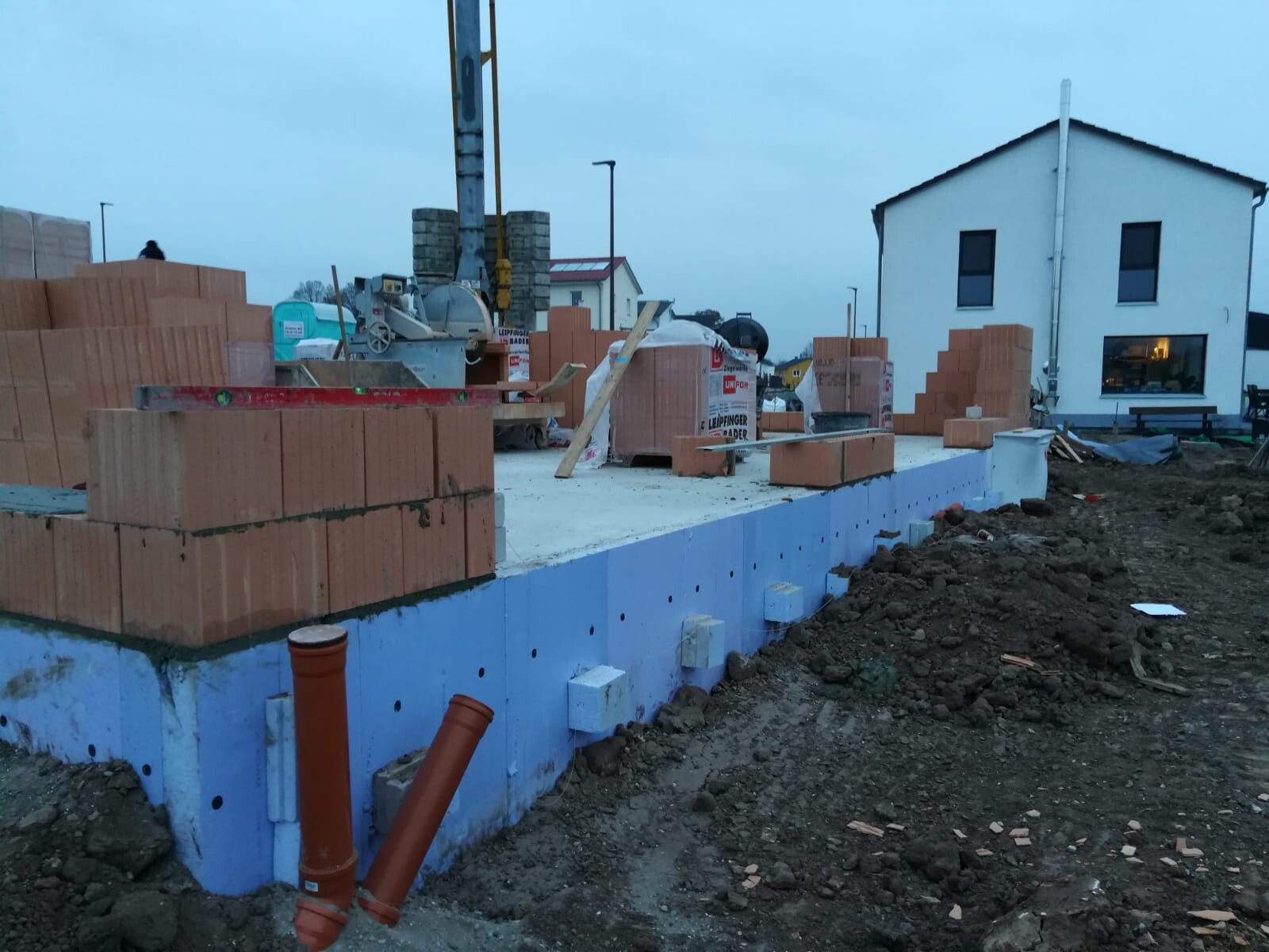 Baustelle mit Backsteinblöcken, Fundamentplatte, Kran und Haus im Hintergrund.