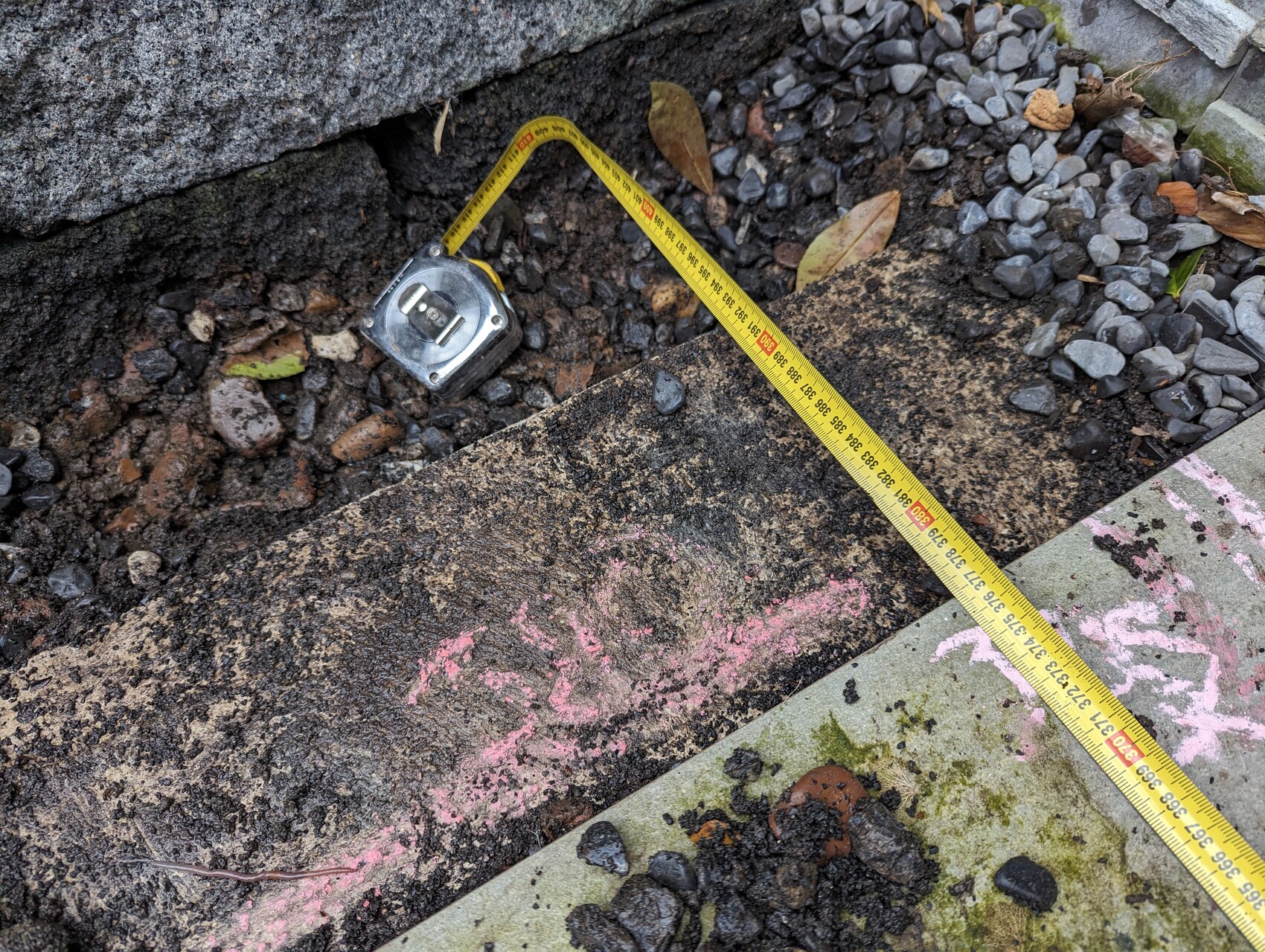 Gelbes Maßband liegt diagonal über Erdaushub, Kies und rosa Markierungen neben einer Betonkante.