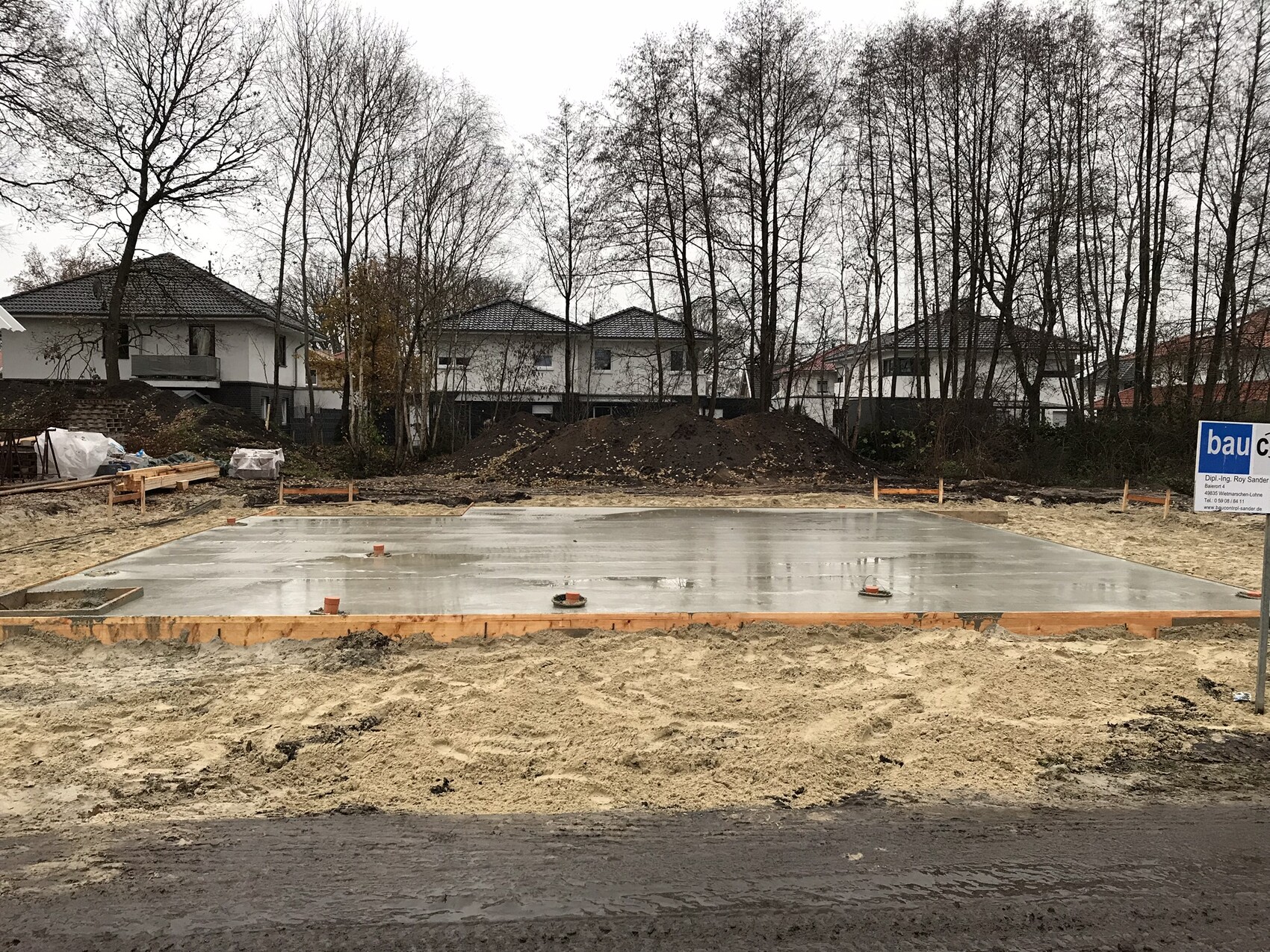 Baustellengrund mit frisch gegossenem Fundament und Holzrahmen, Wohnhäuser im Hintergrund.