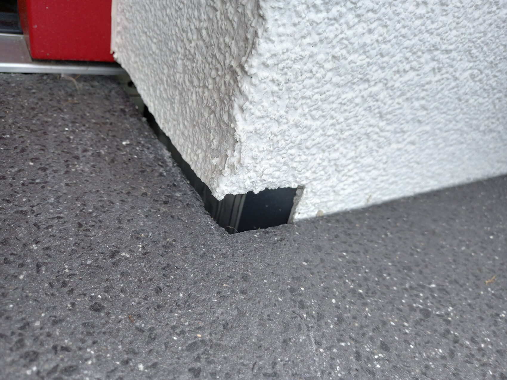Kleines quadratisches Loch im dunklen Asphalt neben rauer weißer Putzwand; dunkler Innenraum.