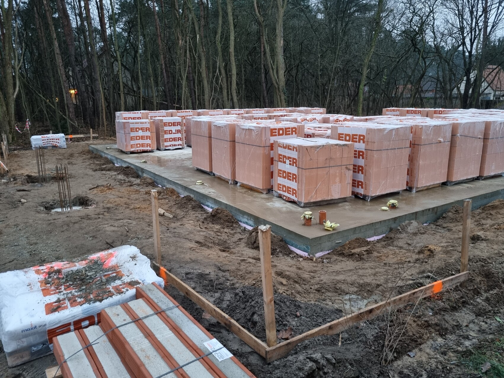 Baustelle: Betonfundament mit gestapelten EDER-Ziegelpaletten, Wald im Hintergrund.