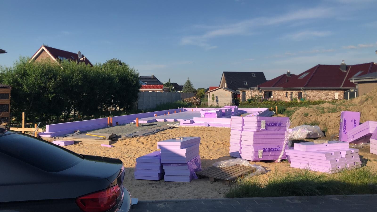 Baustelle mit gestapelten lilafarbenen Dämmplatten, Sandboden und angrenzenden Wohnhäusern.