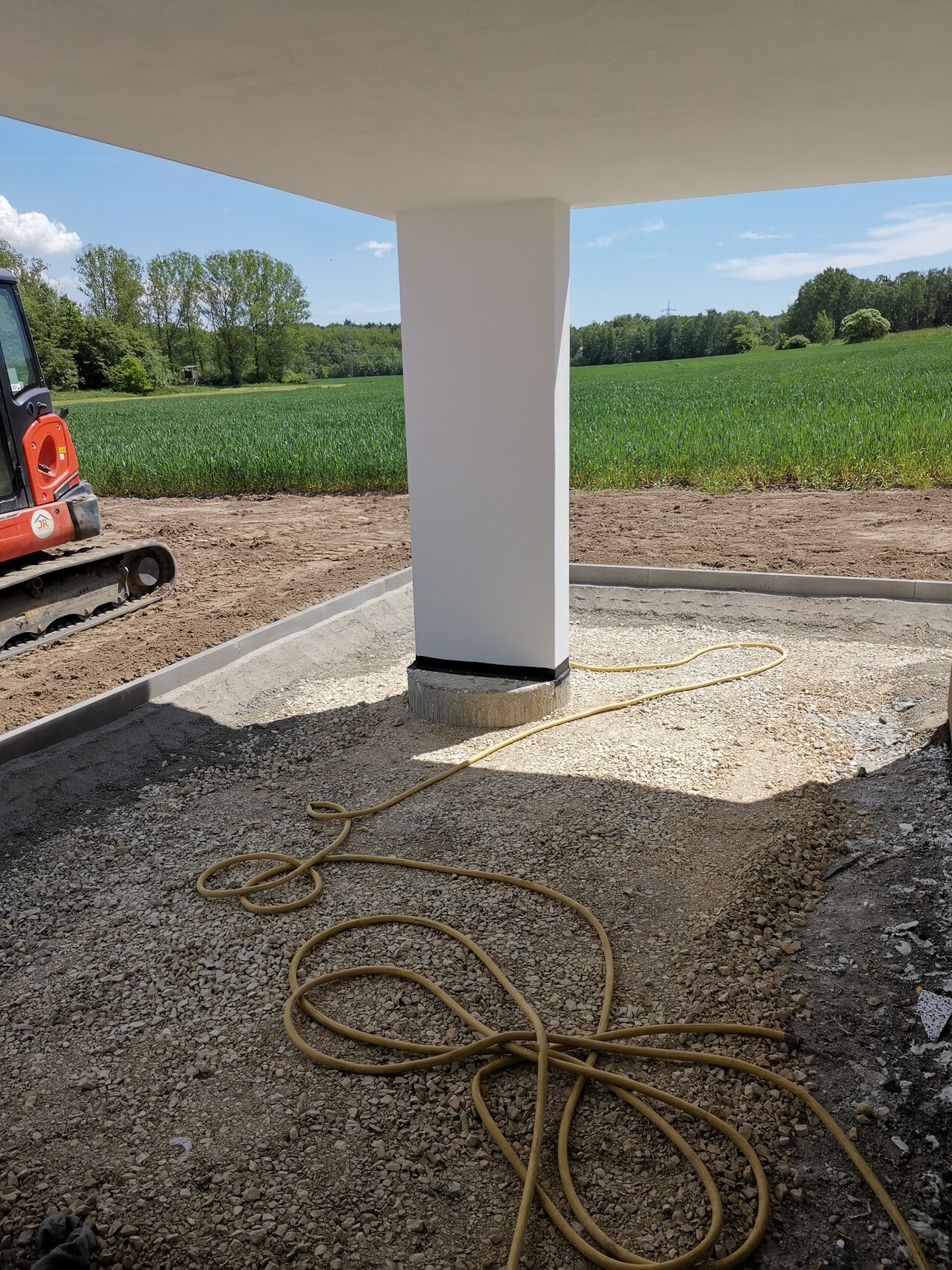 Weiße Betonstütze unter Überbau, Baugrund mit Kies, gelbes Kabel liegt am Boden, ländliche Umgebung.