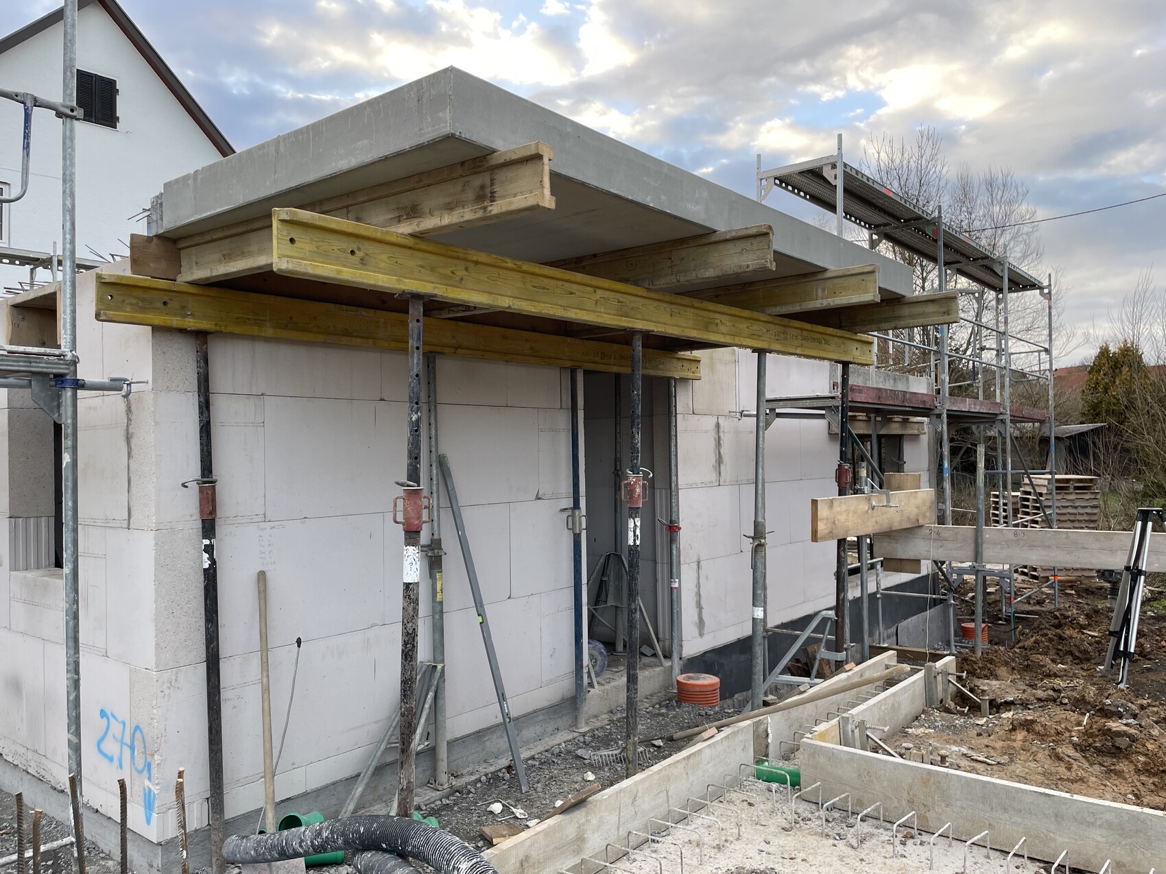Baustelle mit Gerüst, Betonplatte auf Holzbalken über weißem Mauerwerk.