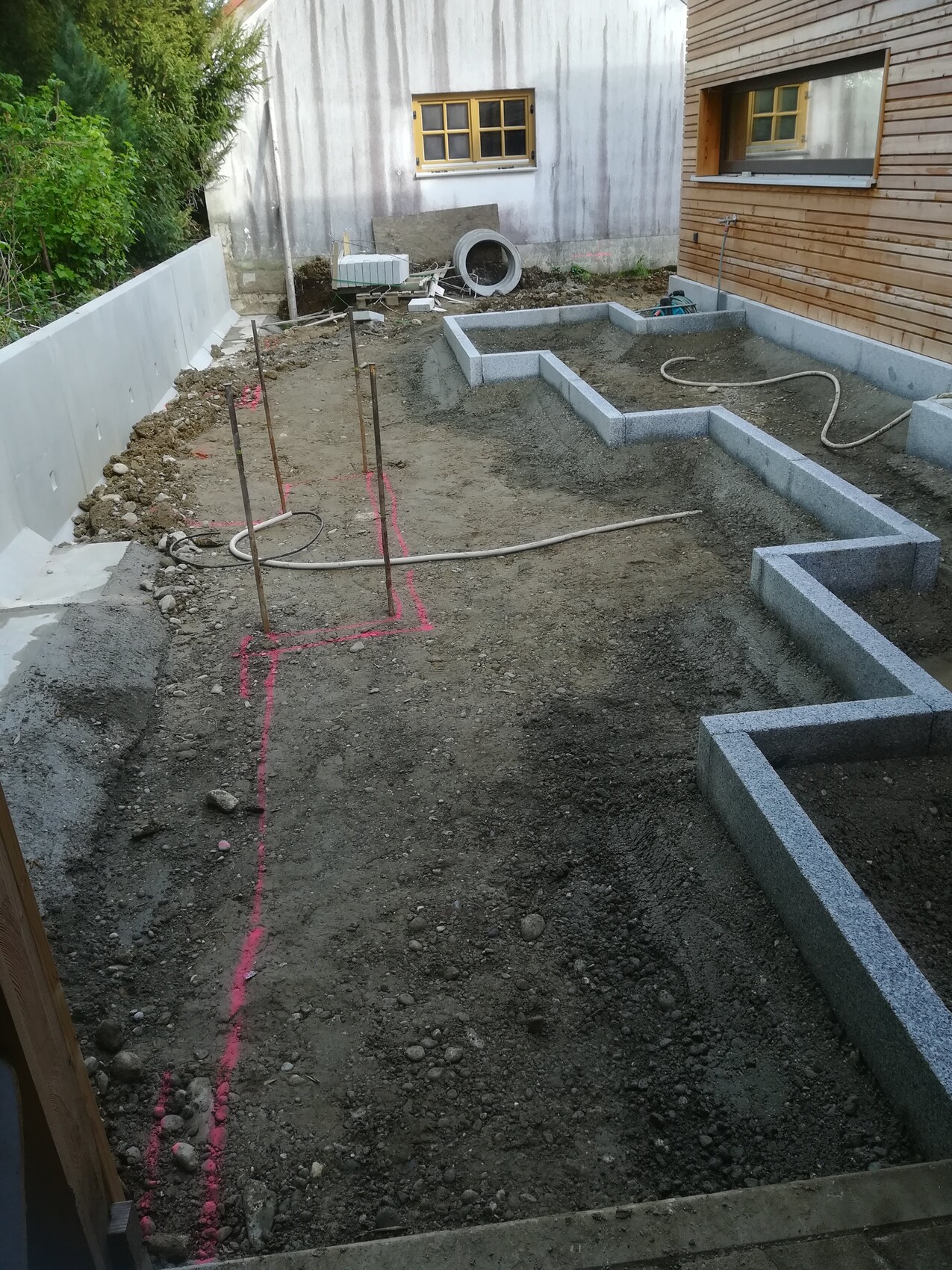 Baustelle mit Fundamentgräben, Stahlstangen, pink markierte Linien und Holzhaus im Hintergrund
