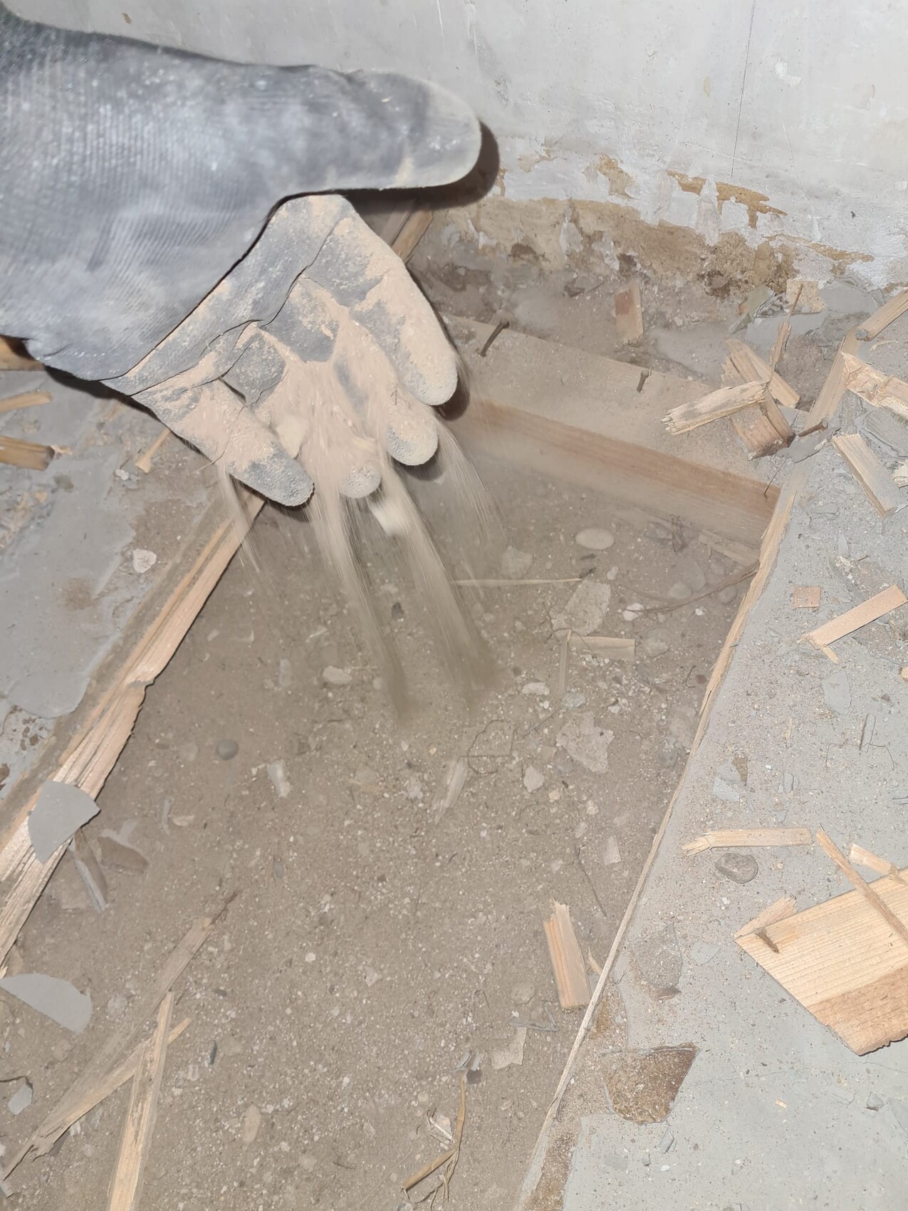 Hand in staubigem Arbeitshandschuh wirbelt Staub aus einem Loch im Betonboden, Holzsplitter liegen daneben.