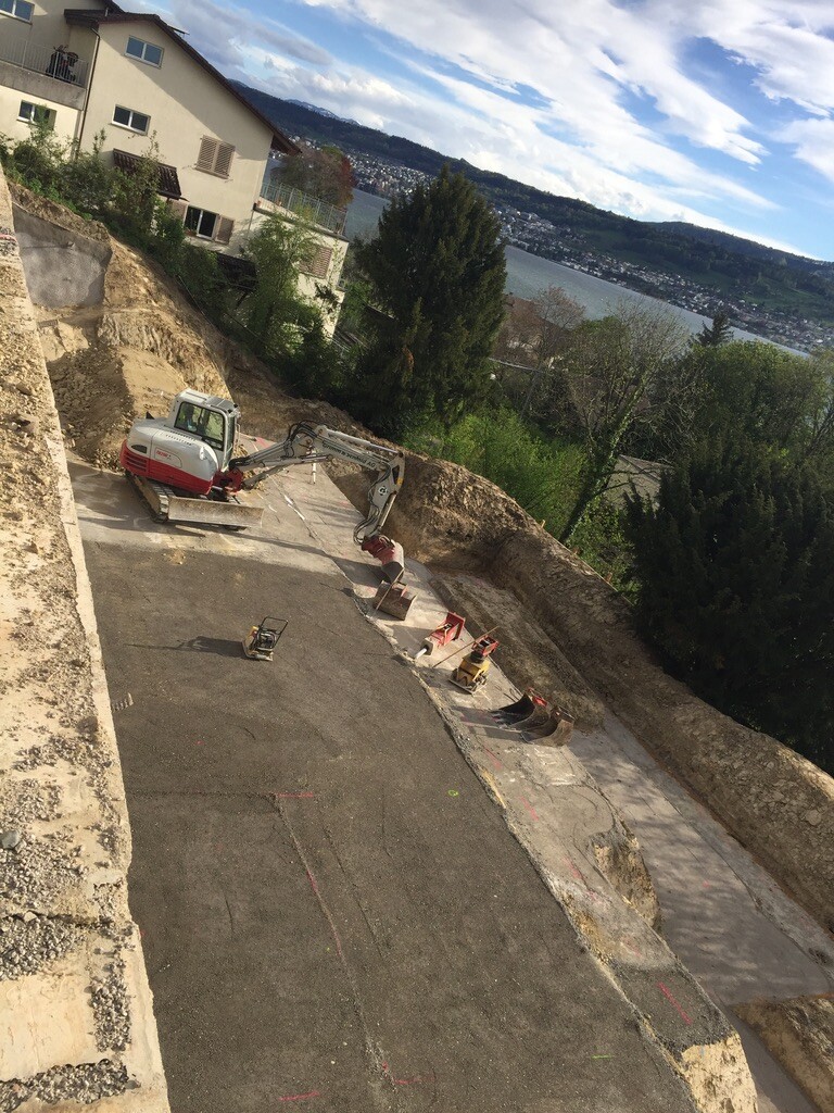 Baustelle an Hang mit Bagger, Absperrungen und verlegter Straße, Blick auf See und Berge.