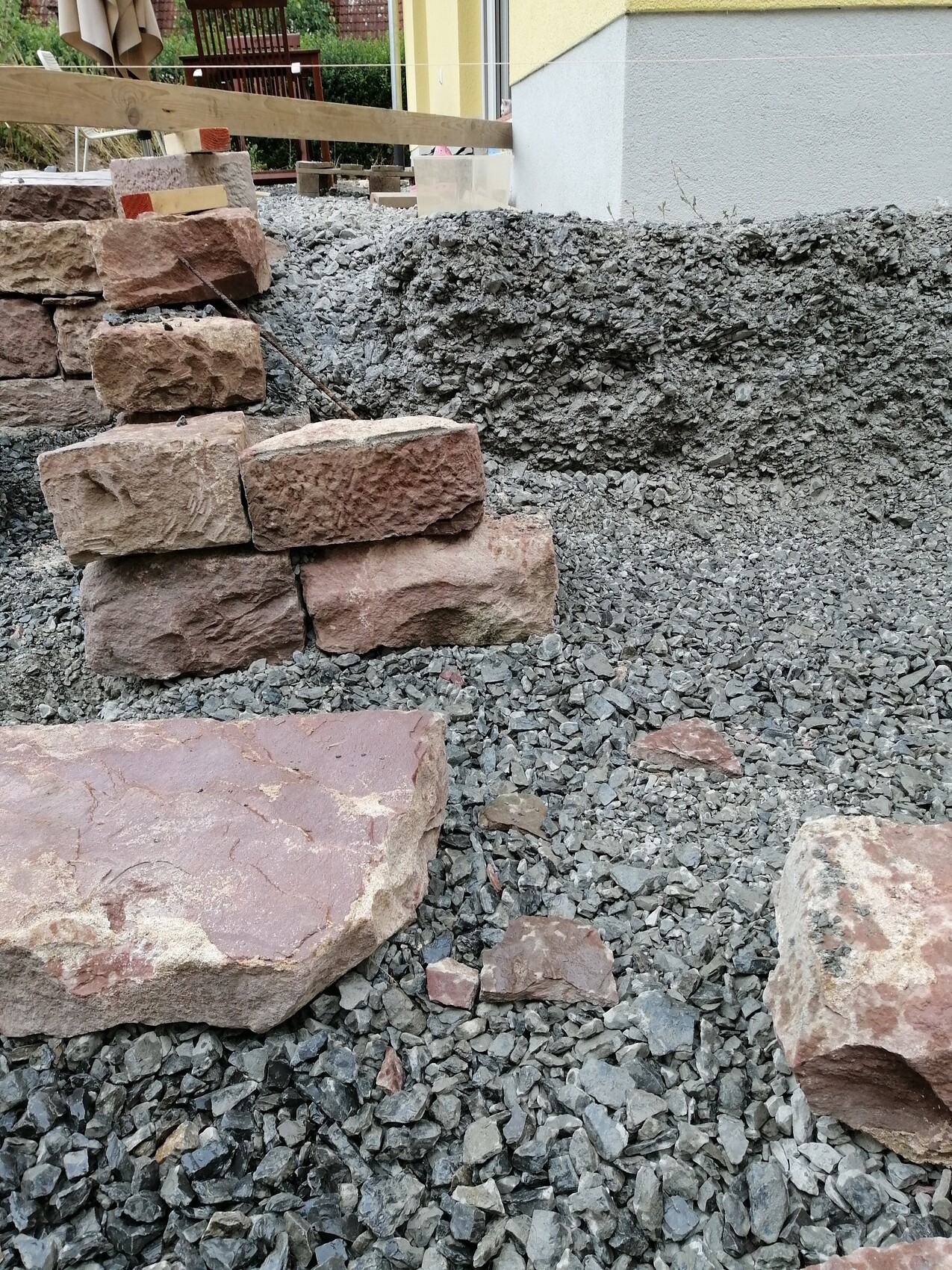 Baustelle: Große bräunliche Steinblöcke als Stufen auf Kies, gelbe Hauswand im Hintergrund