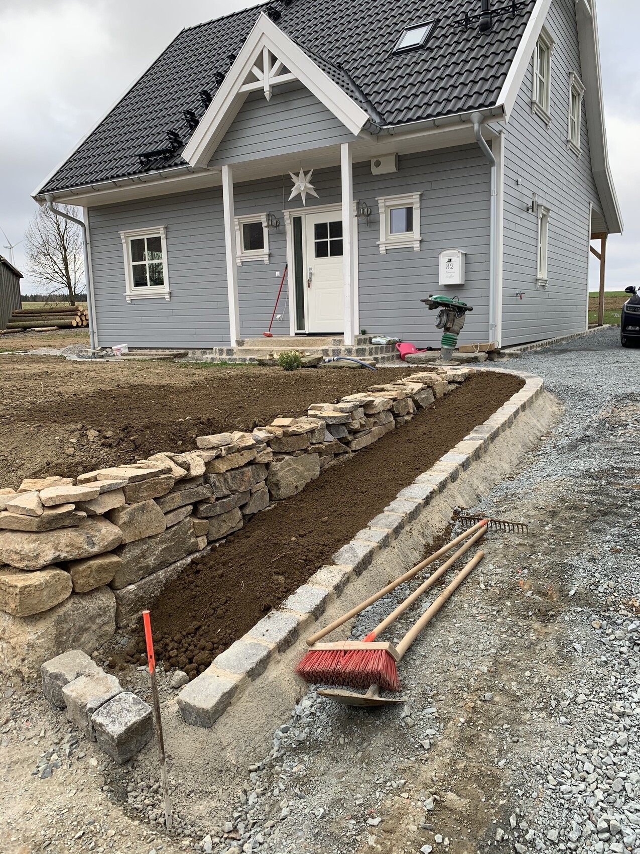 Graues Haus mit Baustelle vor dem Eingang: Steinmauer, Erde und Werkzeuge wie Besen und Rechen.