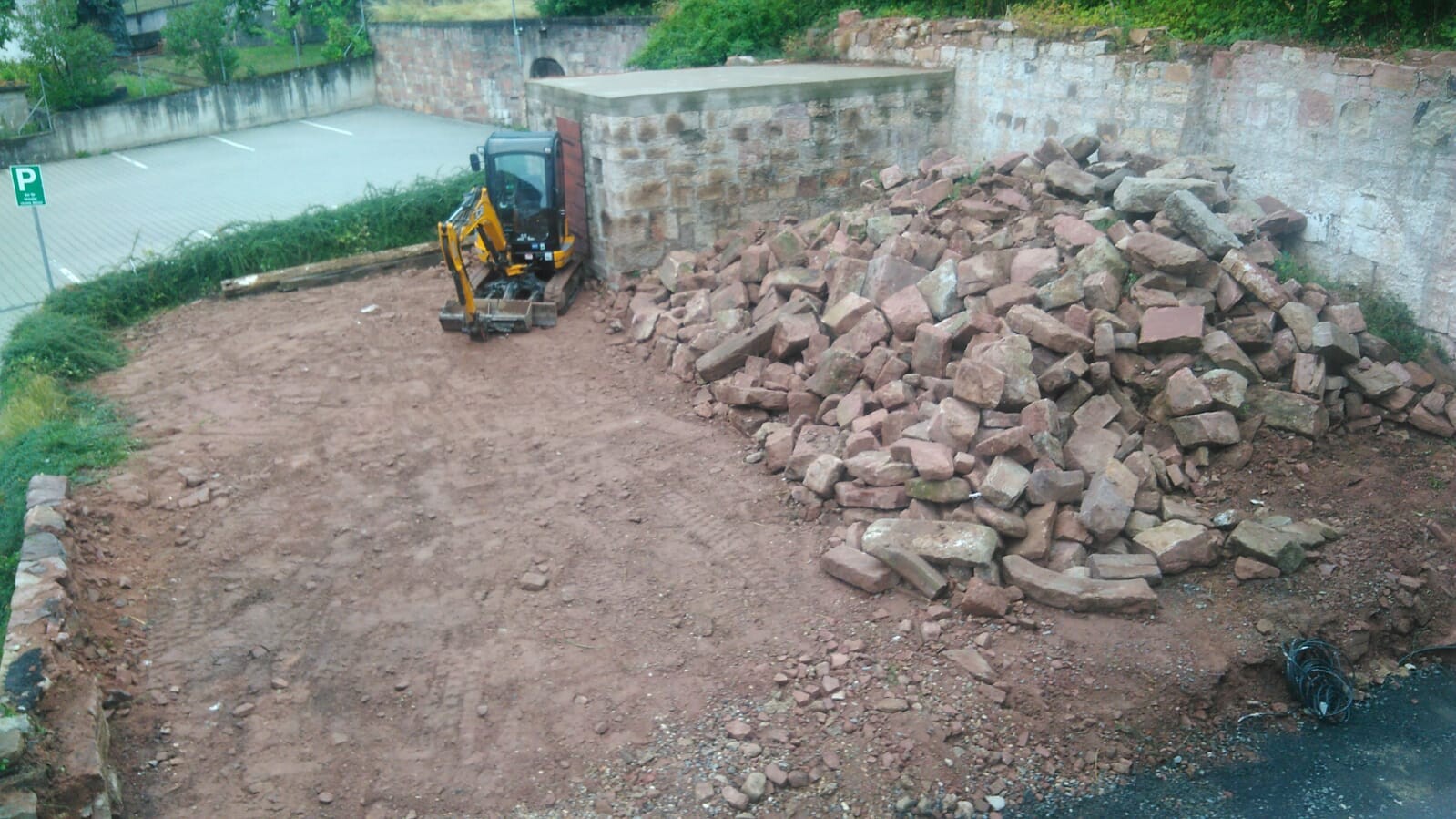 Kleiner Bagger auf einer Baustelle neben Steinmauer und großen Steinhaufen