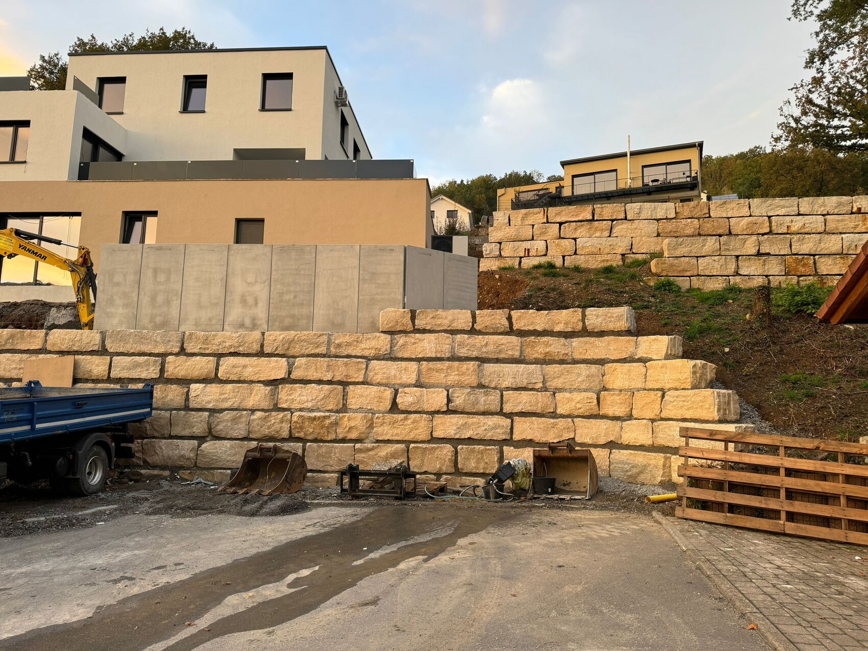 Baustelle mit großen Naturstein-Quaderblöcken als Stützmauer, Bagger und moderne Häuser dahinter.