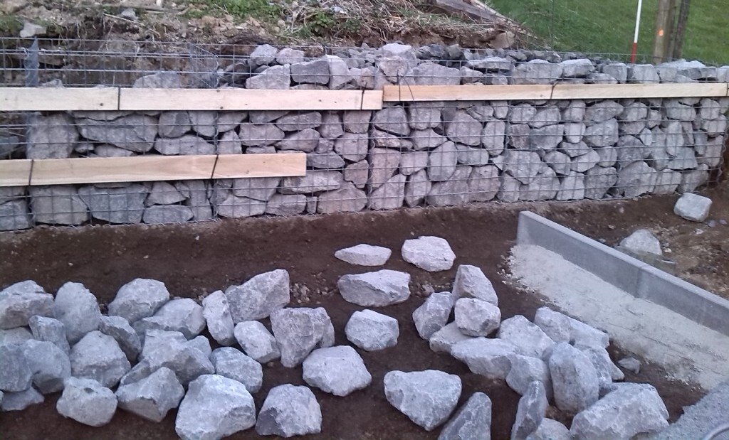 Gabionenwand aus Steinblöcken mit Holzbrettern darüber; lose Steine auf der Baustelle.