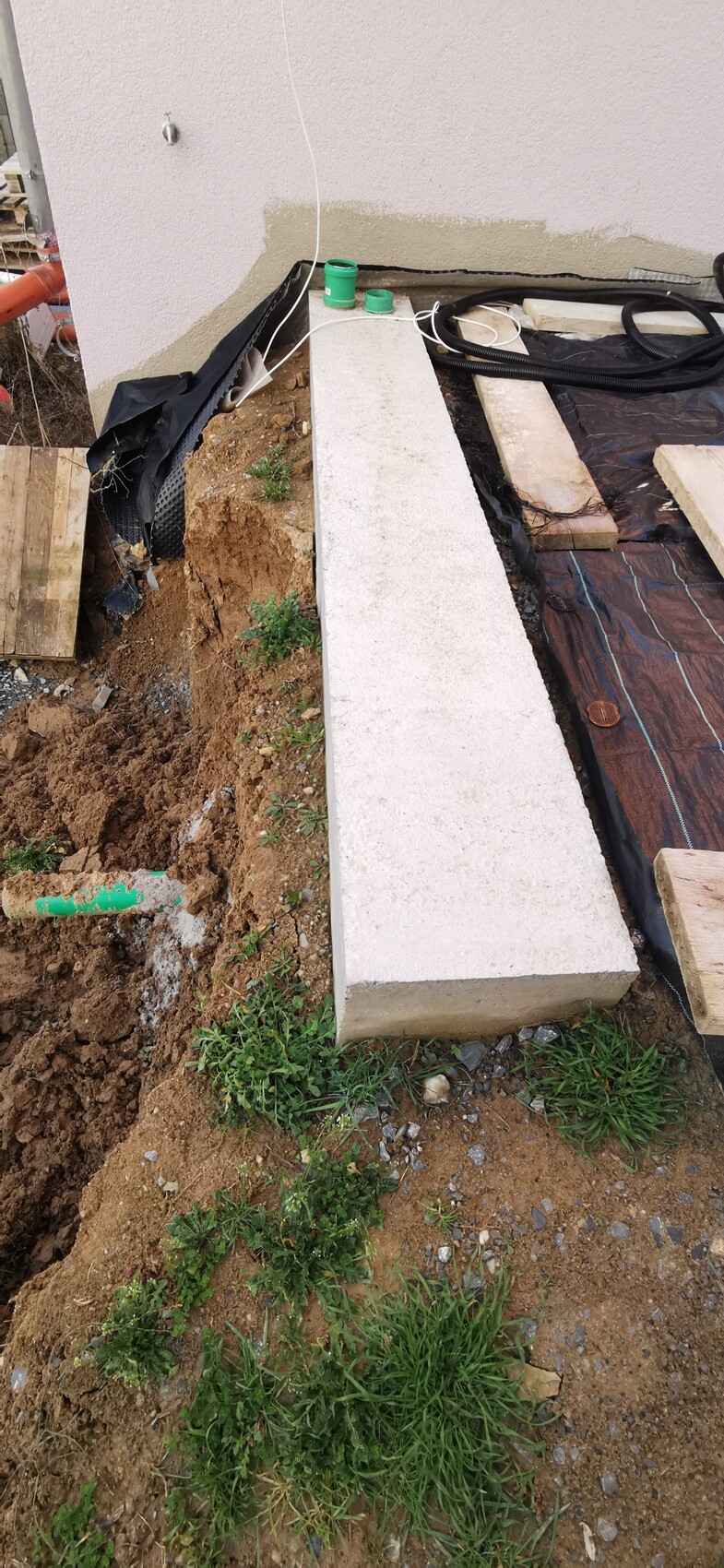 Langer Betonblock neben Außenwand; Erdreich, Rohre, Plastikfolie und Holzreste.