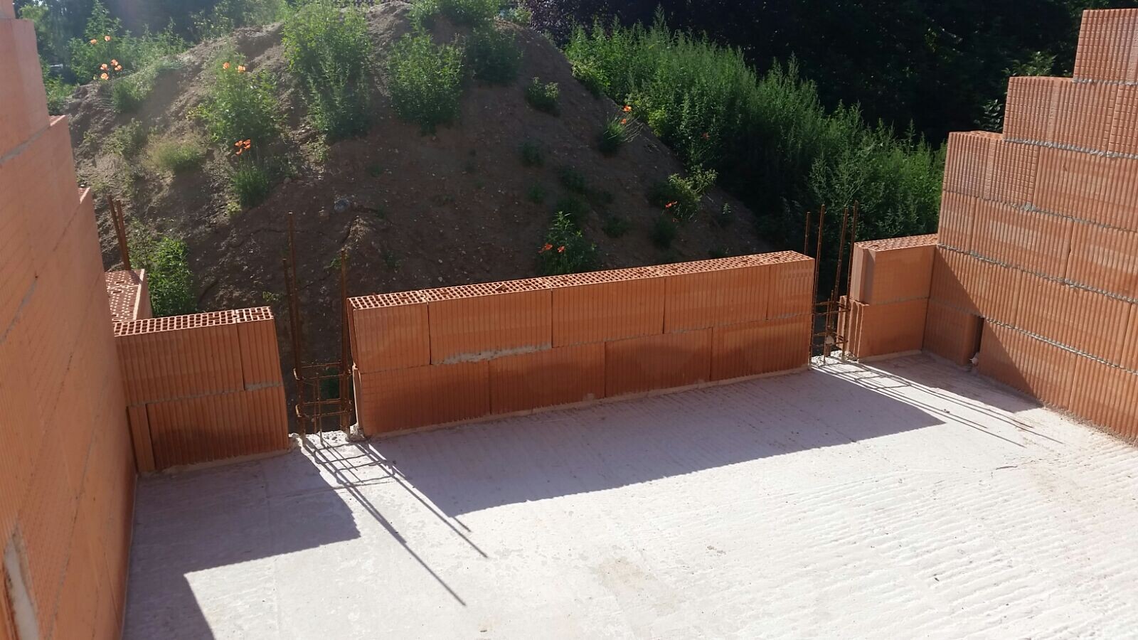 Rohbaustelle: orange Ziegelwände, freigelegte Erdfläche dahinter, Metallstangen ragen hervor.