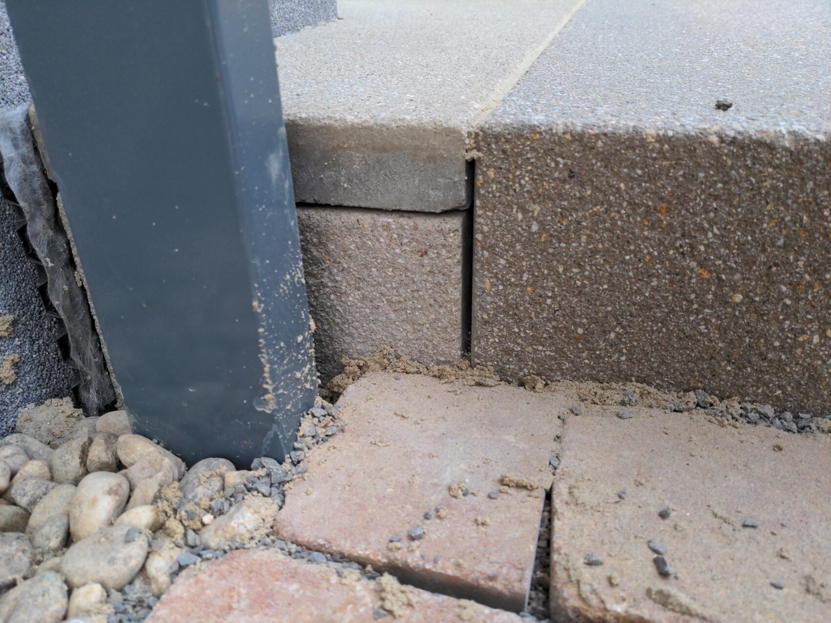 Dunkler Metallpfosten neben einer Mauer aus Beton- und Pflastersteinen mit Sand und Kies.