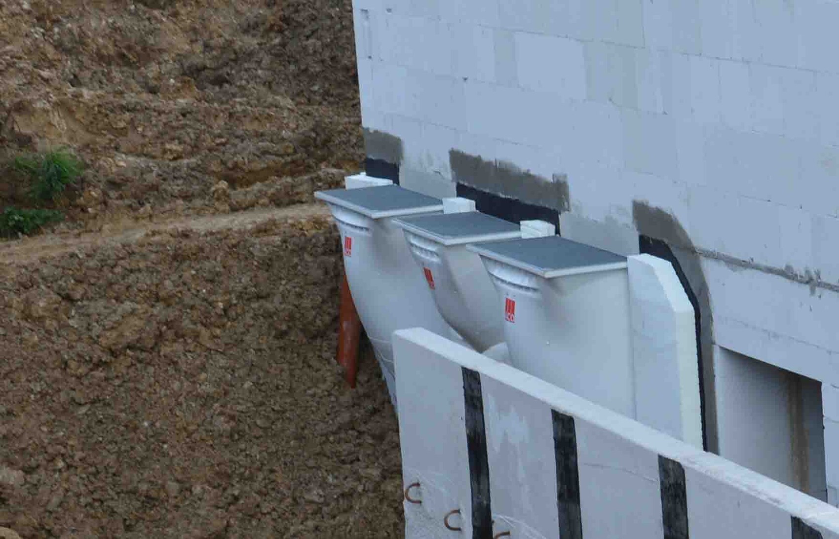 Außenansicht eines Rohbaus mit Fundamentwand und drei Abfallcontainern auf schlammigem Gelände