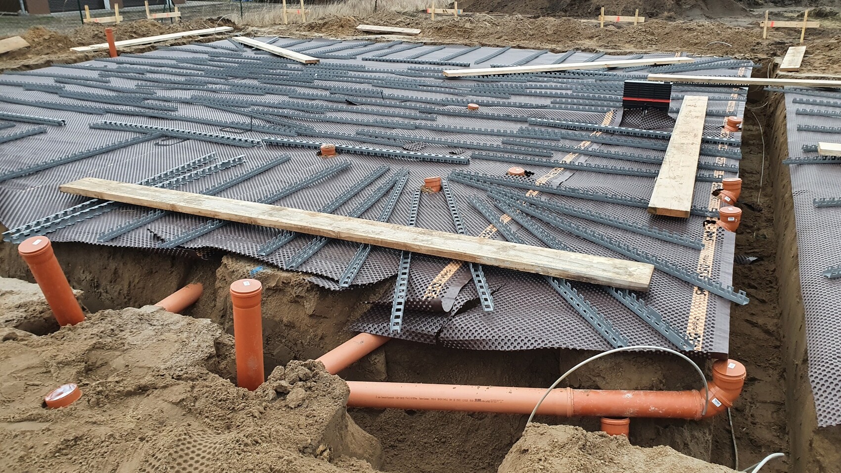 Baustelle: graue perforierte Bodenmatte, orange PVC-Rohre und Holzbohlen sichtbar.