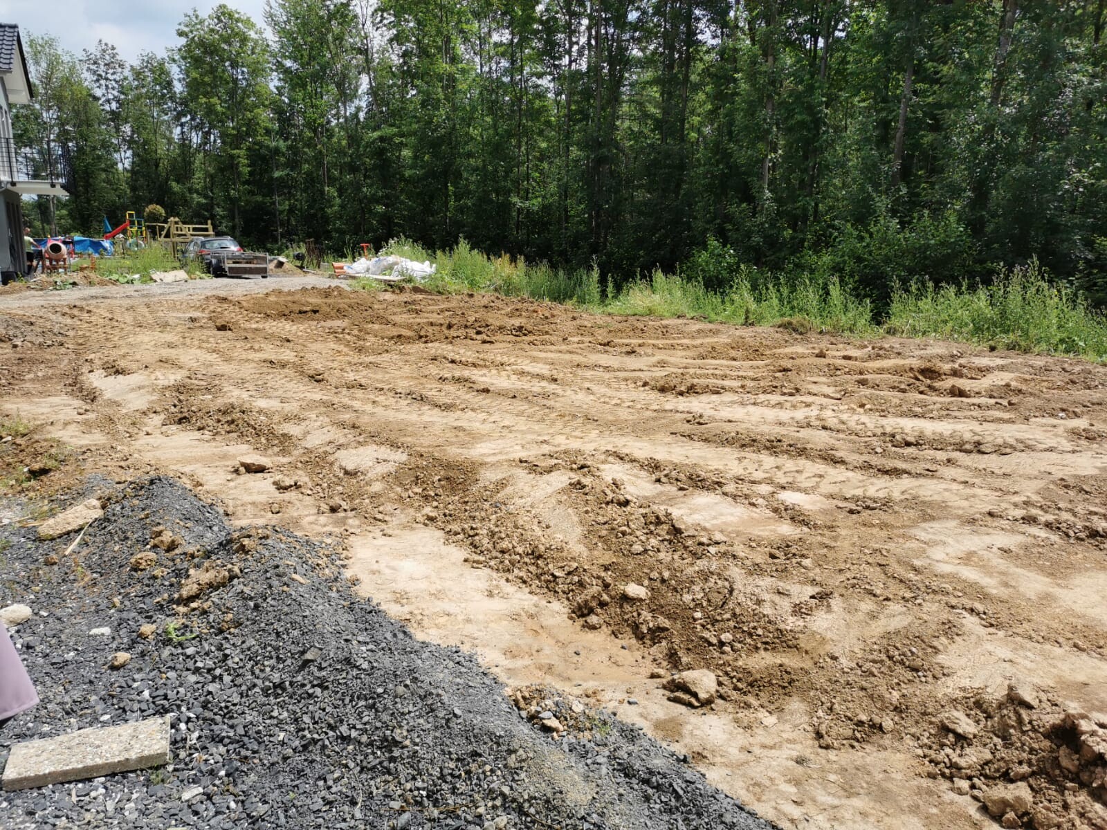 Baustelle im Freien mit aufgeschütteter Erde, Kieshaufen und Wald am Rand.