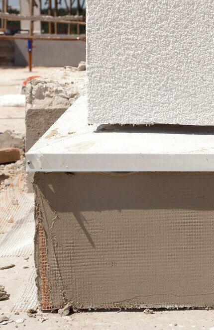 Nahaufnahme eines Rohbaus: Betonblock auf Styroporfundament, Baugerüste im Hintergrund.