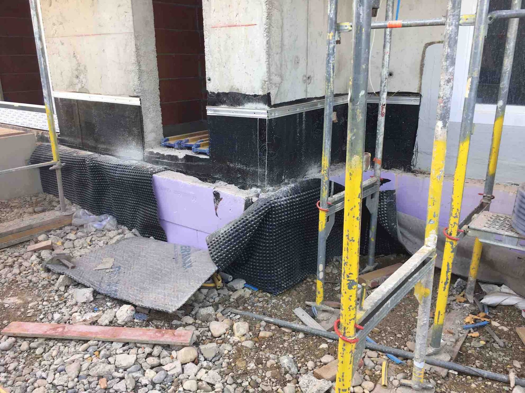 Baustelle mit Fundament, Wandbau, Gerüst, Dämmplatten und Kiesboden.