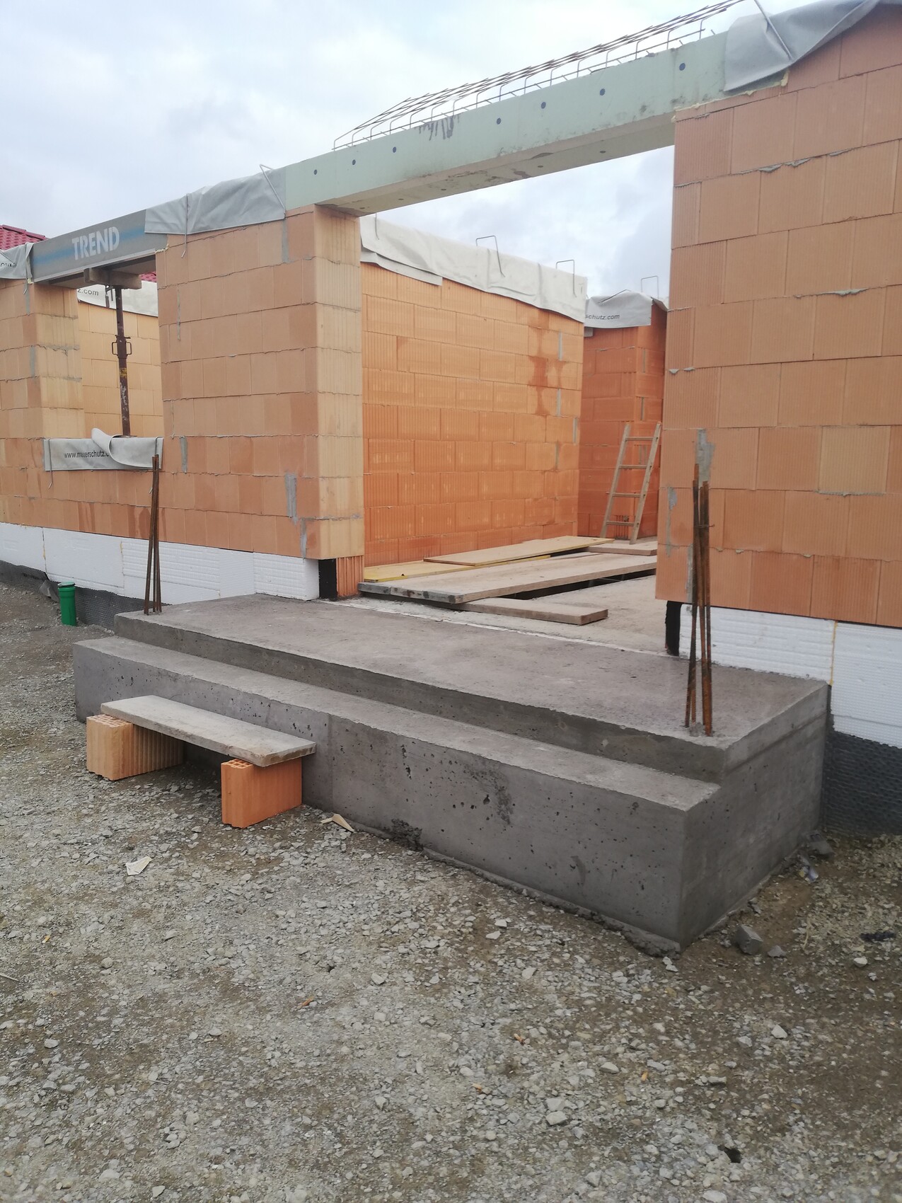 Baustelle mit orange Ziegelwänden, Betonstufen, Leiter und Holzplanken im Eingangsbereich.
