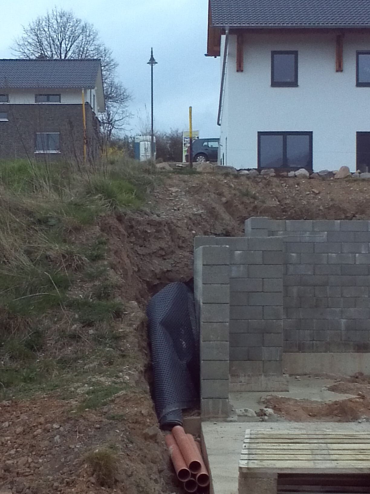 Baustelle: Hang mit Erdreich, schwarze Drainageröhre neben Blockwand; Haus im Hintergrund.