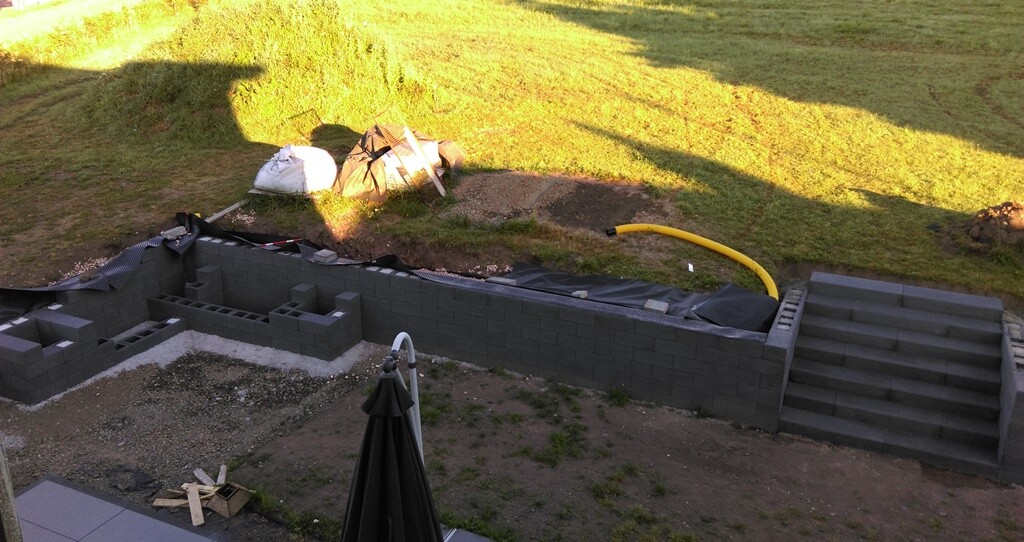 Gartenbau-Baustelle mit Fundamentmauern aus Blöcken, Treppen, gelber Schlauch und Baumaterialien.