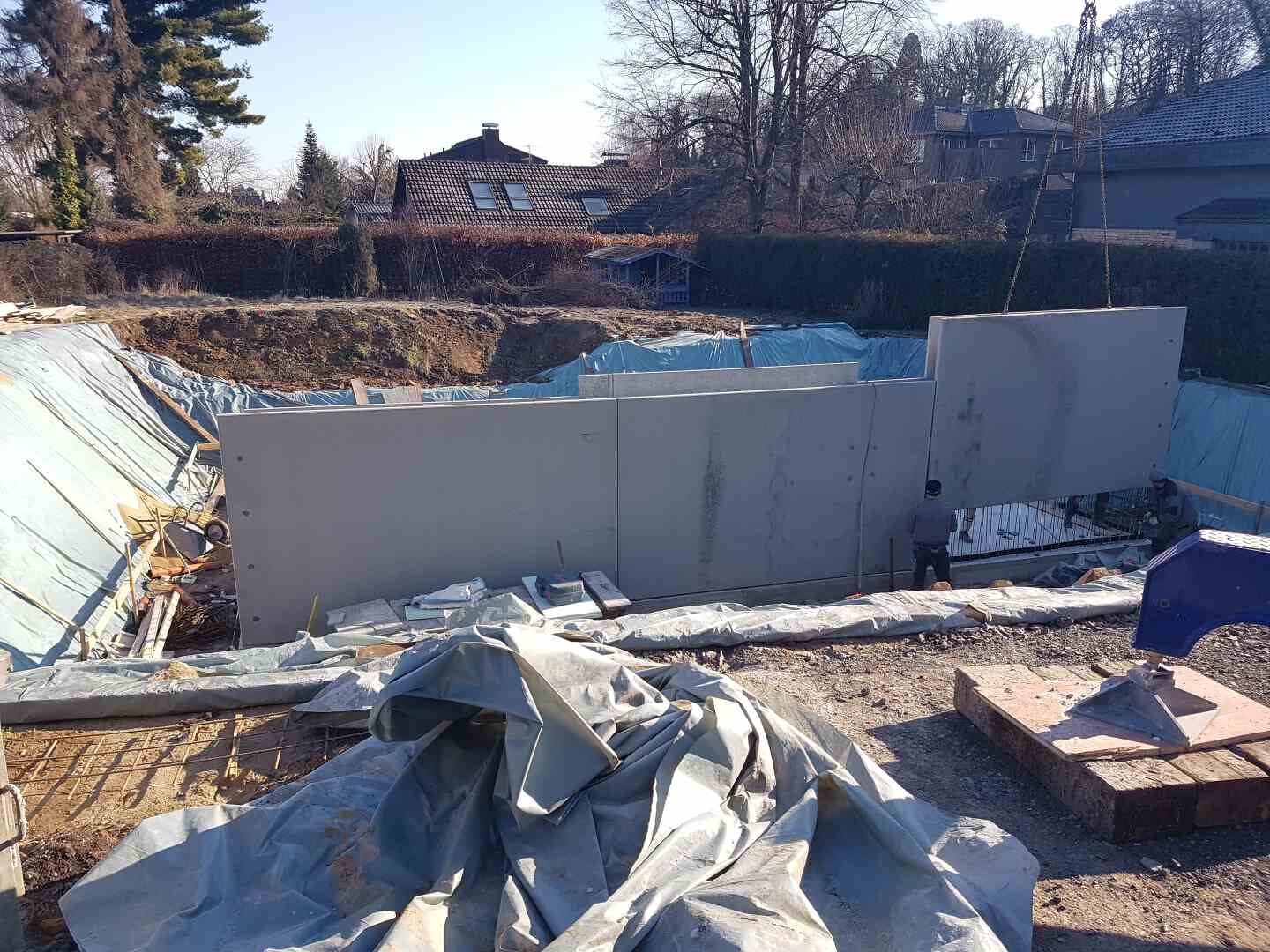Baugrube mit großen Betonplatten, Bauarbeiter und blaue Planen im Hintergrund.